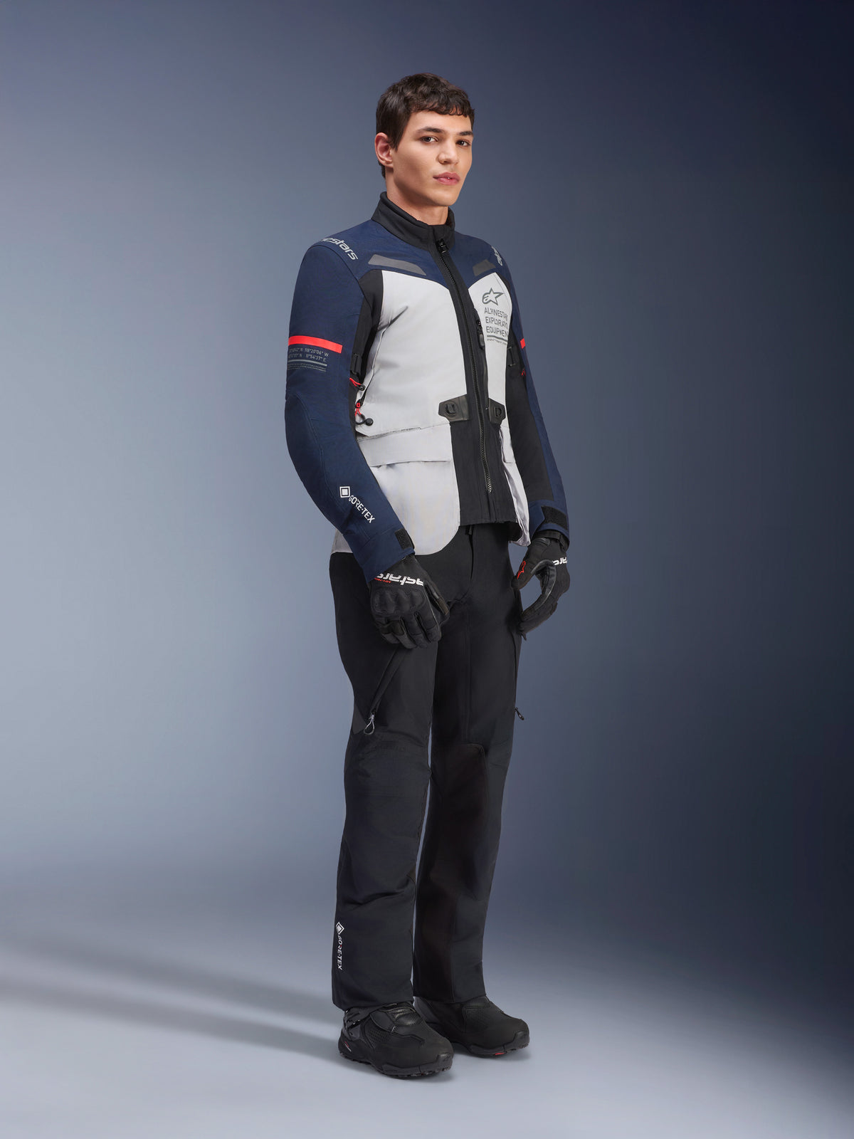 Alpinestars ST-7 2L GORE-TEX-jakke i farvekombinationen Ice Gray, Dark Blue og sort med røde detaljer, båret af en mandlig model med sorte motorcykelbukser og handsker, professionelt adventure touring-motorcykeludstyr