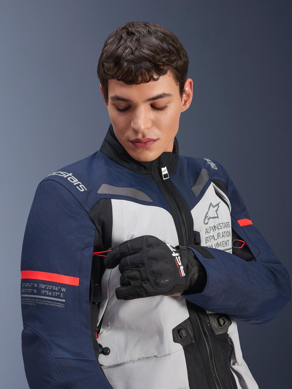 Alpinestars ST-7 2L GORE-TEX jakke, Ice Gray, mørkeblå og sort med orange detaljer, bæres af en mand, der justerer en detalje på ærmet, med 'Alpinestars Exploration Equipment' branding og geografiske koordinater, professionelt adventure-motorcykeludstyr