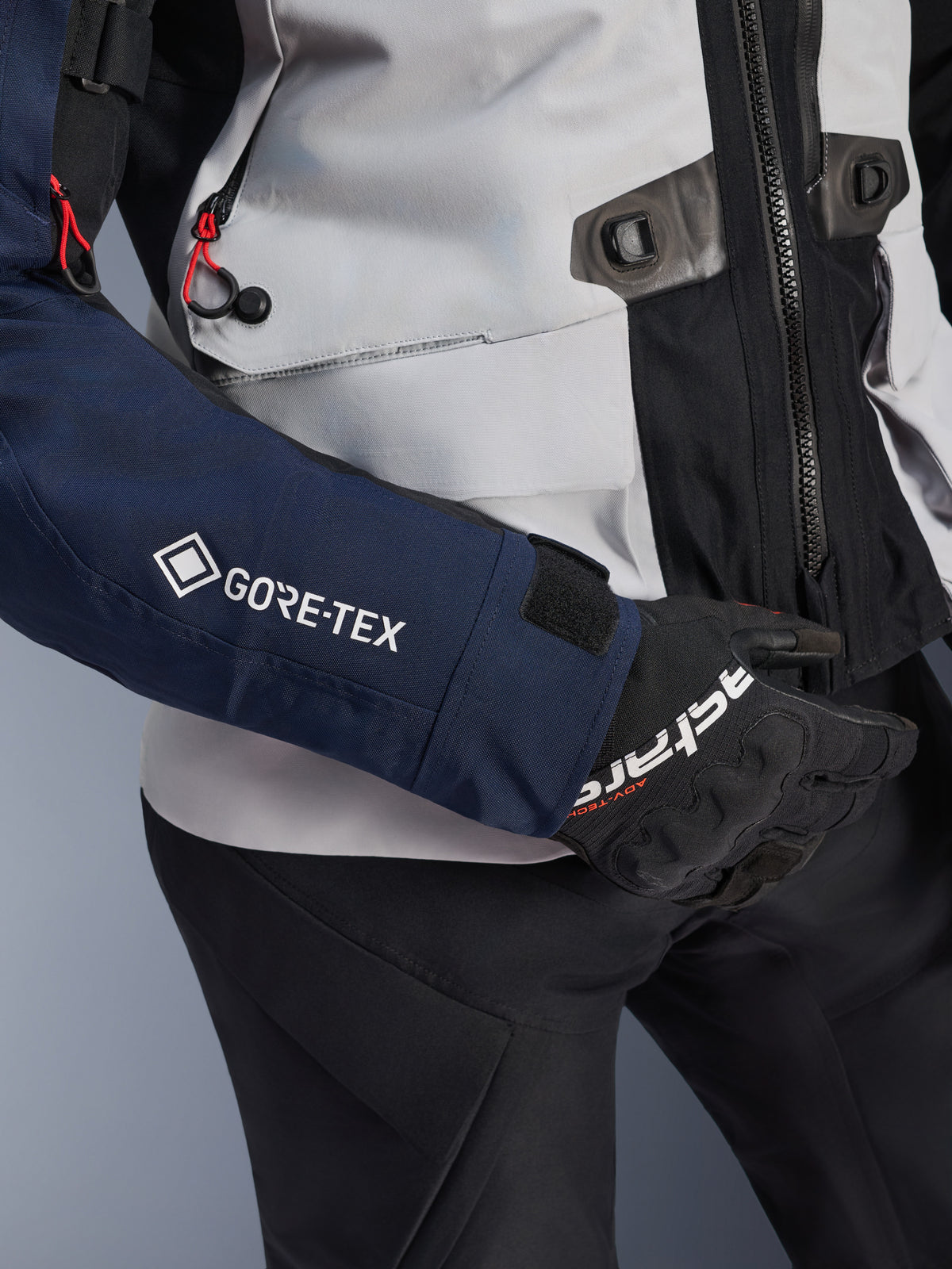 Alpinestars ST-7 2L GORE-TEX jakke, nærbillede af mørkeblåt ærme med hvidt GORE-TEX-logo, isgrå torso med sorte detaljer og røde snoreaccenter, båret med sorte Alpinestars-handsker og -bukser, adventure touring-motorcykeludstyr