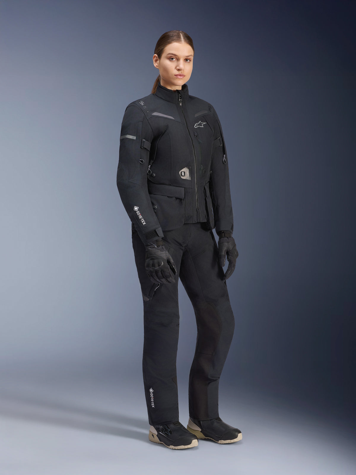 Kvindelig model iført Alpinestars Stella ST-7 2L GORE-TEX adventure touring-sæt, sort og mørkegrå jakke og bukser, med tydelig GORE-TEX-branding på ærmet og benet, kombineret med sorte handsker og kørestøvler, professionelt motorcykeludstyr designet til touring i al slags vejr.