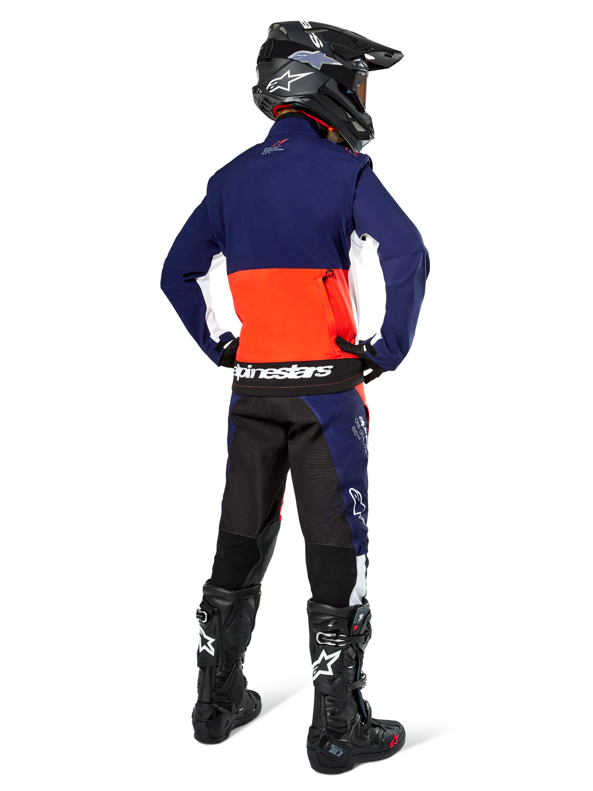 Motocross rider from the back wearing Alpinestars Lite-Dura Softshell Jacket in Navy, Hot Orange, and White, showing the large rear storage pocket. Styled with matching enduro pants, a black motocross helmet, and black off-road boots.