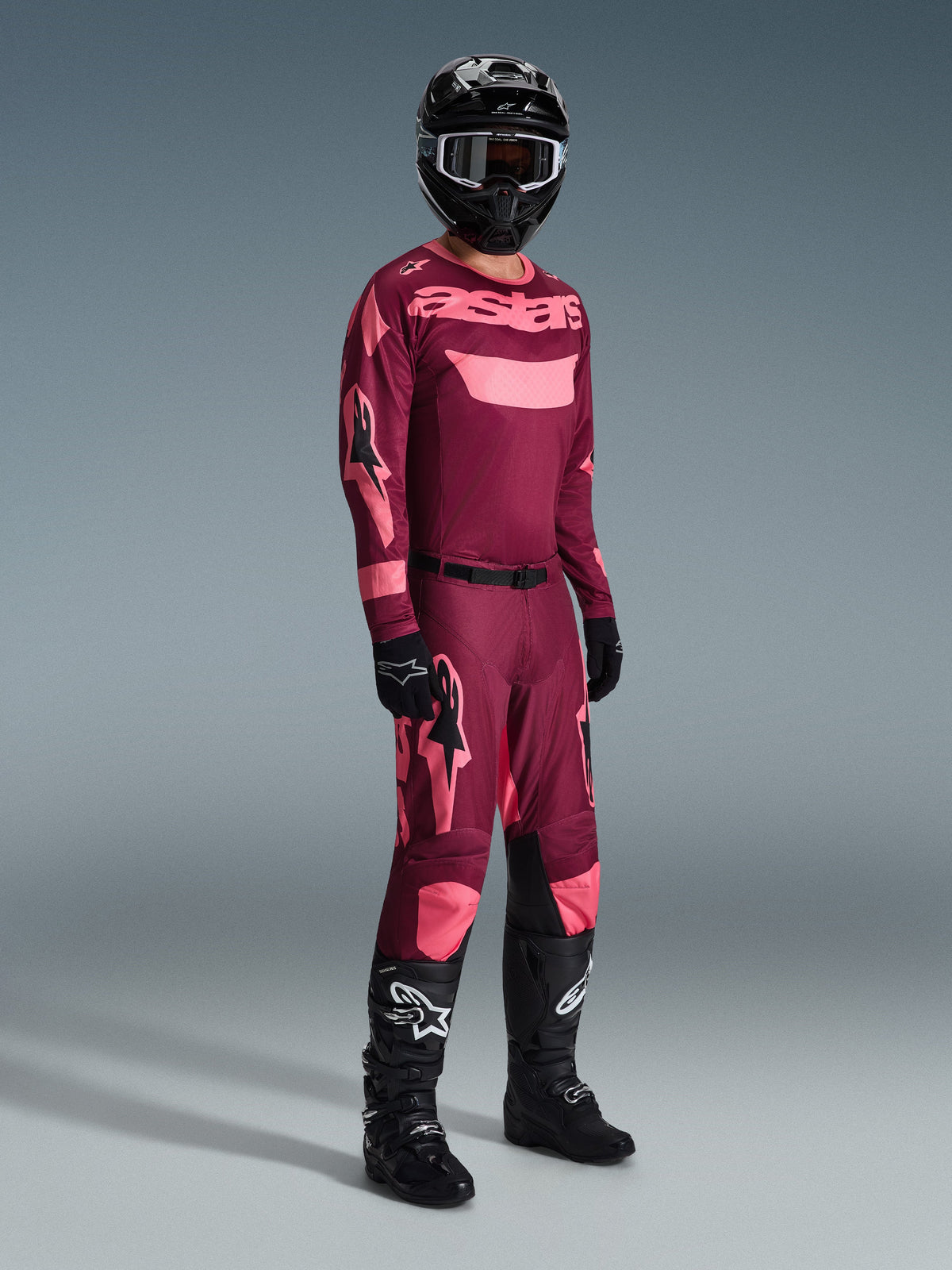 Motocross-kører iført komplet racerudstyr, Alpinestars 2026 Racer Riway trøje i mørkerød koral med pink detaljer, matchende mørkerøde bukser med pink logoer, sort hjelm med hvidkantede goggles, sorte handsker og sorte støvler med hvide logoer, koordineret outfit til off-road kørsel