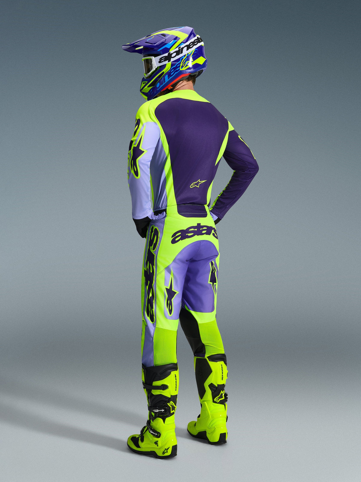 Motocross-kører set bagfra iført Alpinestars 2026 Racer Portl-bukser i Purple Yellow Fluo, stylet med en matchende lilla og gul trøje, hjelm og knaldgule racerstøvler, med markant 'astars'-branding på bagdelen og stjernelogoer på benene.