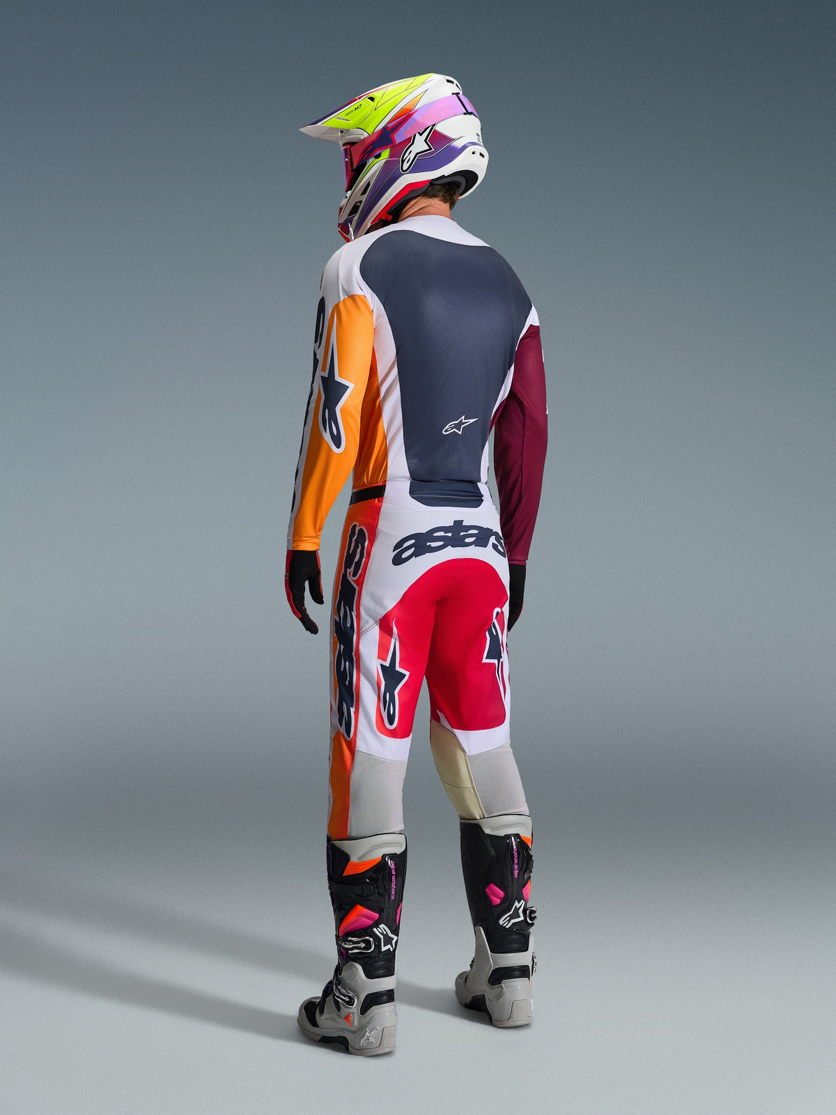 Motocrosskører set bagfra iført komplet racerudstyr, Alpinestars 2026 Racer Portl bukser i rød, lysegrå og mørkegrå, matchende trøje med orange og bordeauxrøde ærmer, flerfarvet grafisk hjelm, grå og sorte off-road støvler med lyserøde detaljer, professionelt motocross-outfit