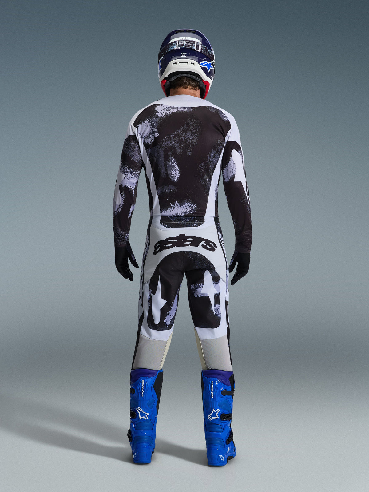 Motocrosskører set bagfra iført Alpinestars 2026 Racer Lahnd-trøje og matchende bukser i Iron Camo med sort-hvidt splatter-mønster, kombineret med klare blå støvler og en koordinerende hjelm, komplet udstyrssæt til off-road racing