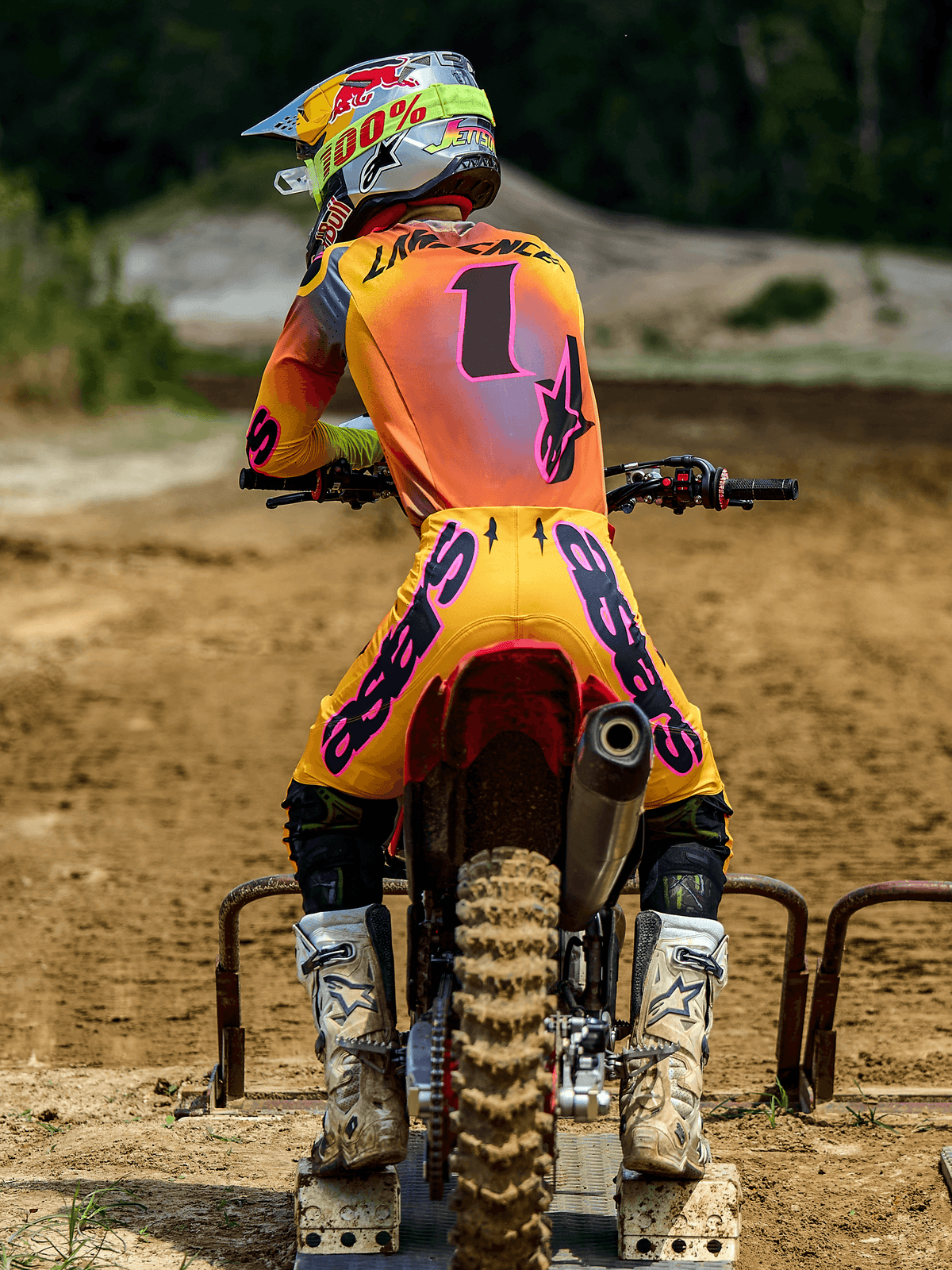 Motocrossrytter set bagfra iført Alpinestars 2025 Supertech Lipan-udstyrssæt, gul og pink, trøje med nummer 1 og 'LAWRENCE' på ryggen, gule bukser med pink 'ASTARS'-branding, hvide støvler og matchende hjelm, siddende på en motocrossmaskine ved startbommen
