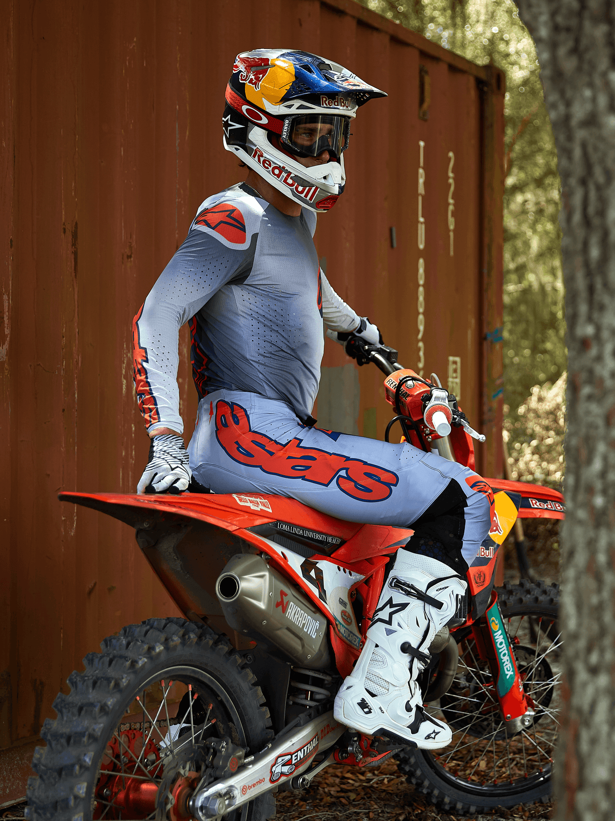 Motocrossrytter siddende på en rød crosser, iført Alpinestars 2025 Supertech Lipan-udstyrssæt i Gray Orange Fluo, lyseblå bukser med markant orange Astars-logo, matchende jersey, hvide motocross-støvler og en Red Bull-hjelm, koordineret professionelt racerudstyr