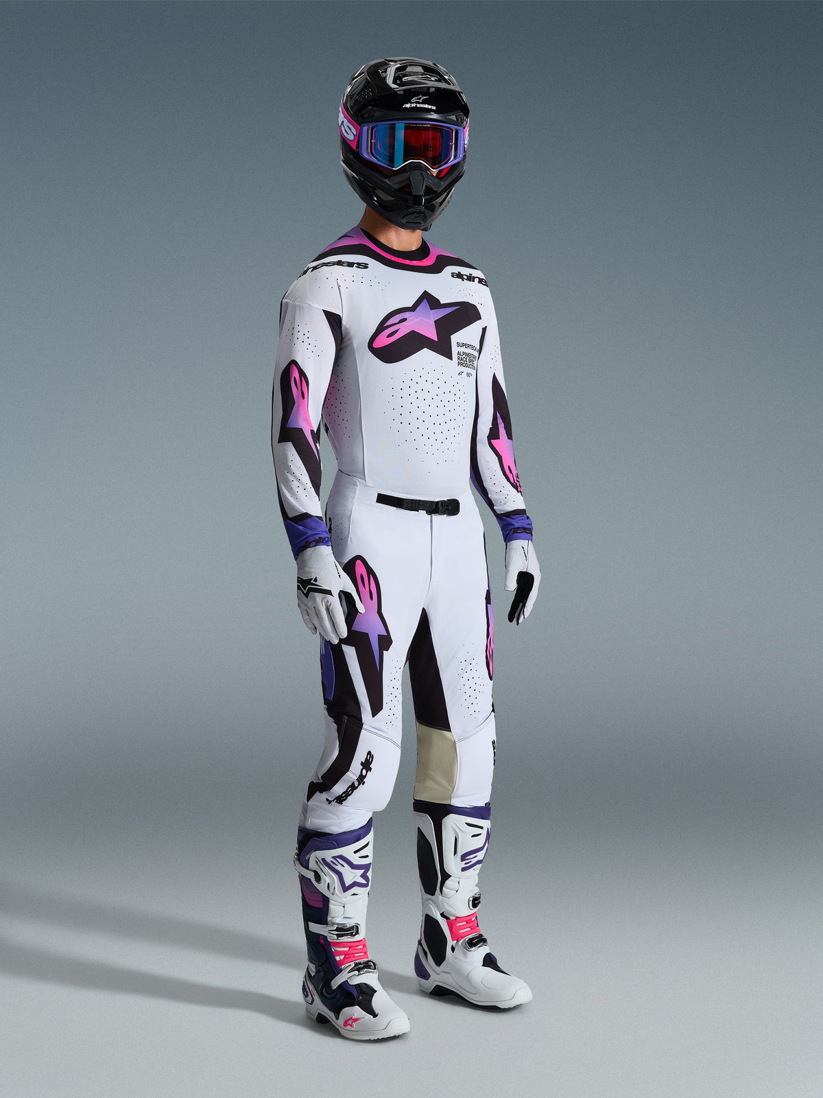 Motocross-kører iført et komplet Alpinestars racersæt med 2026 Supertech AFD Vista bukser i hvid, lilla og sort, matchende trøje, sort hjelm med lilla goggles, hvide og lilla støvler samt hvide handsker, koordineret professionelt terrænkørselsudstyr.