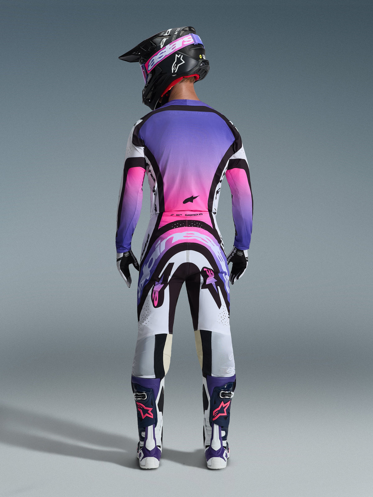 Motocrosskører set bagfra iført komplet racerudstyr, lilla og pink trøje med gradient, Alpinestars 2026 Supertech AFD Vista bukser i hvid, lilla og sort, sort hjelm med lilla goggles, hvide og lilla støvler, koordineret off-road racersæt.