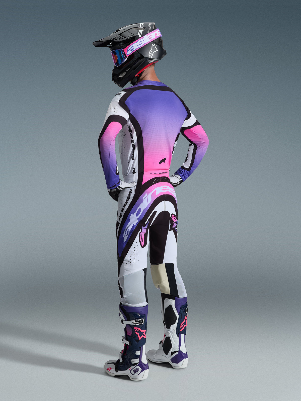 Motocrosskører set bagfra iført Alpinestars 2026 Supertech AFD Vista bukser, hvid, lilla og sort farvekombination, kombineret med en matchende lilla og pink gradient-trøje, sort hjelm og hvide og lilla racerstøvler for et komplet off-road racing-look.