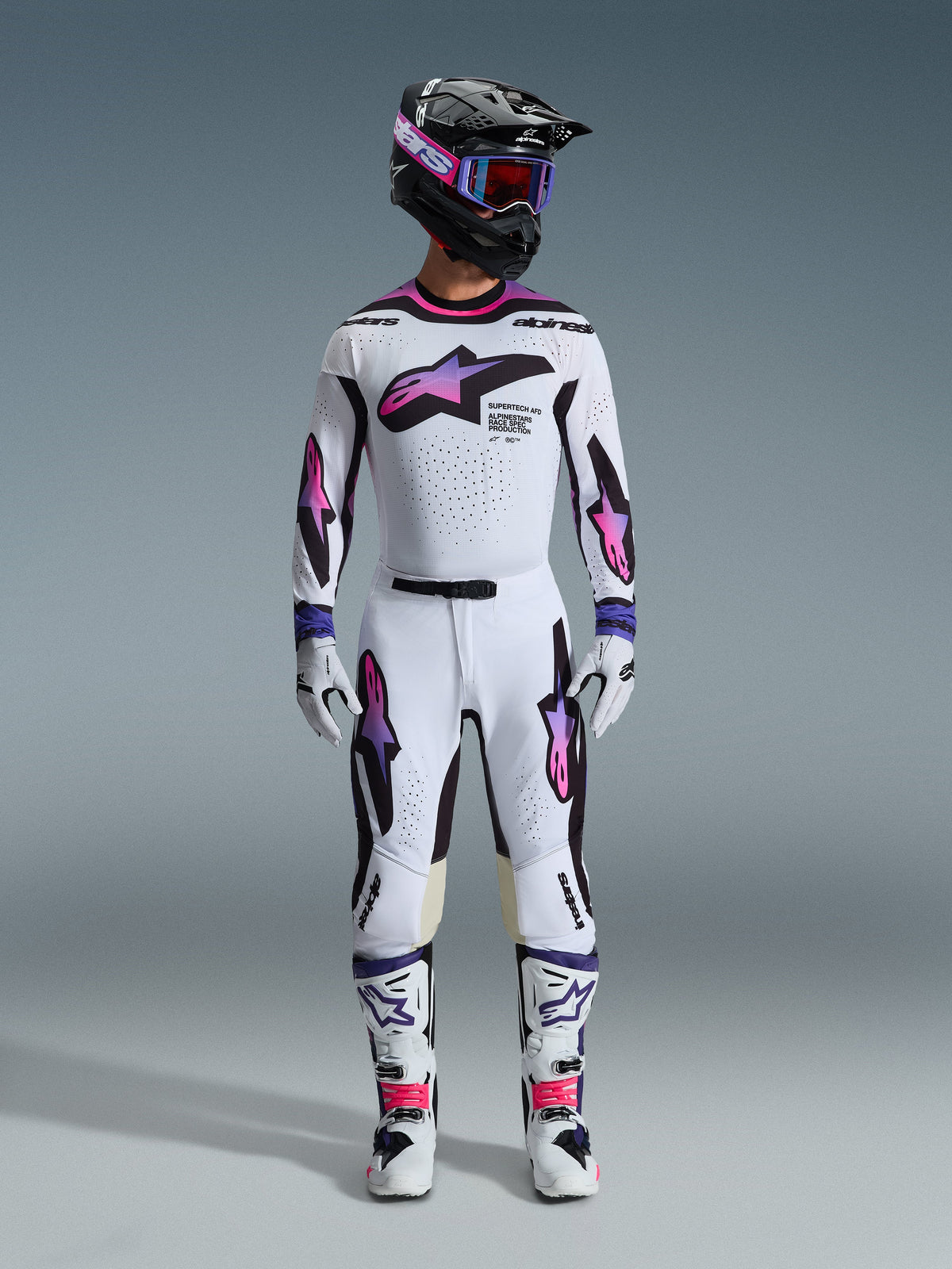 Motocrosskører iført et komplet racersæt, Alpinestars 2026 Supertech AFD Vista bukser og trøje i hvid, lilla og sort, matchende handsker, hvide og sorte støvler, sort hjelm med lilla goggles, slank atletisk pasform til terrænkørsel