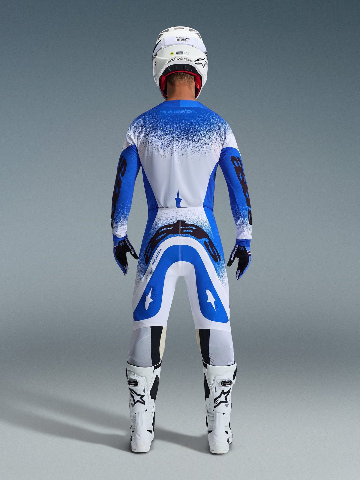 Motocrosskører set bagfra iført komplet racerudstyr, Alpinestars 2026 Supertech Scenz bukser i Ucla Blue og hvid, matchende blå og hvid trøje med nistret mønster, hvid hjelm, hvide og sorte støvler, koordineret sæt til professionel racing
