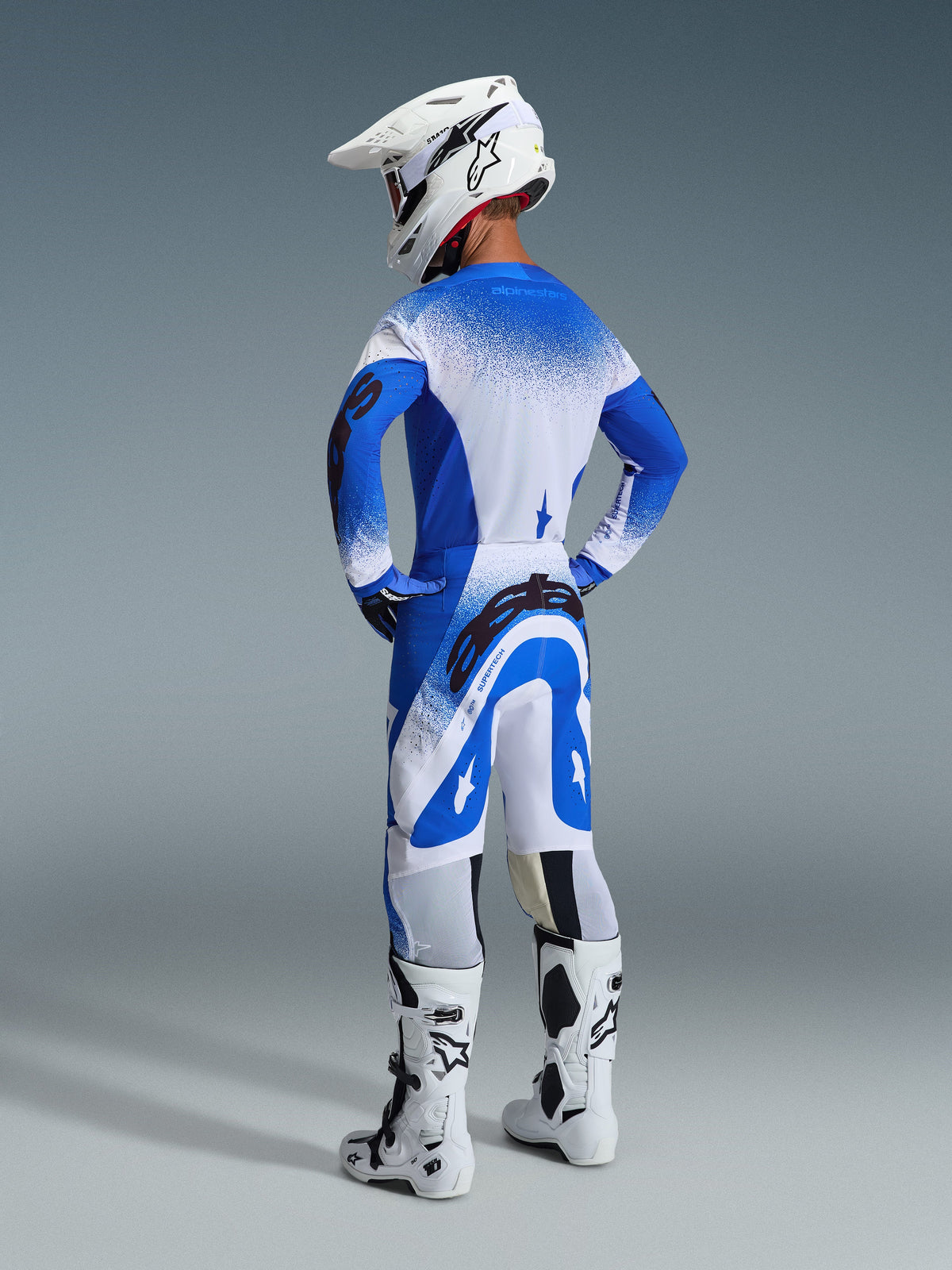Motocrosskører set bagfra iført 2026 Supertech Scenz-bukser og -trøje i Ucla Blue White, hvid hjelm med Alpinestars-logo, hvide og sorte motocross-støvler, professionelt racingudstyrssæt til terrænkørsel.