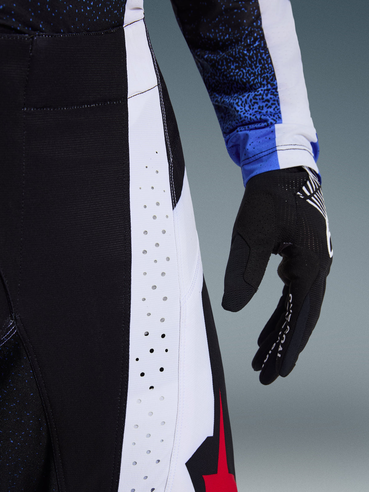 Detaljeret nærbillede af Alpinestars 2026 Techstar Knif bukser i sort, hvid og rød, der viser laserskårne perforationer på et hvidt sidepanel for ventilation, vist med en sort motocross-handske og et blåt mønstret trøjeærme, hvilket fremhæver den højtydende racerkonstruktion.