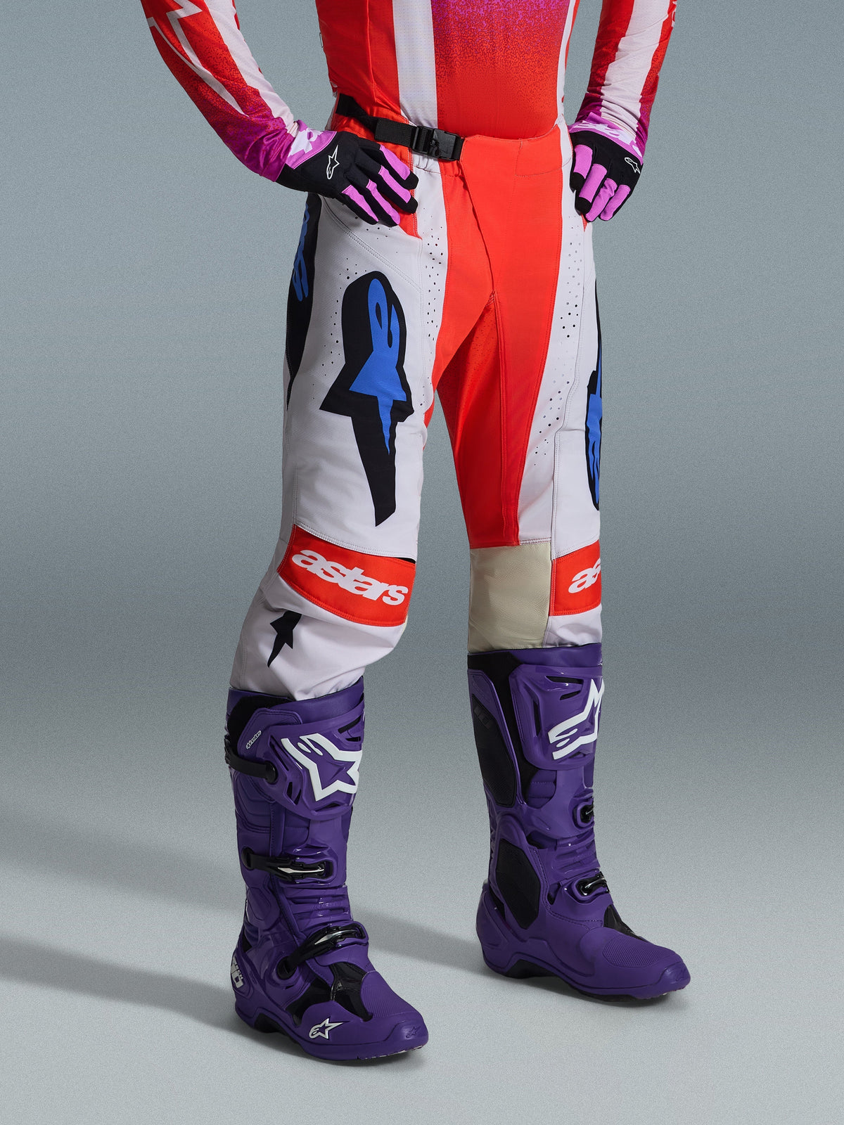 Alpinestars 2026 Techstar Knif bukser, motocross-bukser, hvid og klar rød med blå grafiske accenter, med et 'astars'-logo på knæet, båret med lilla støvler og sorte og pink handsker, professionelt off-road racing-udstyr
