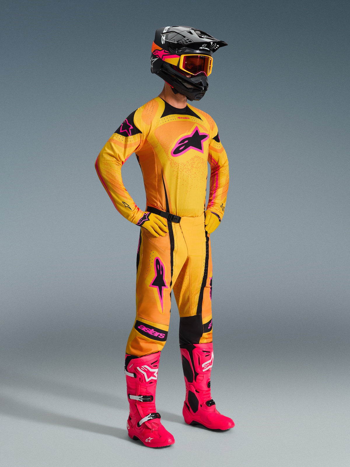 Motocrosskører iført Alpinestars 2026 Techstar Nomur-bukser i orange, gul og pink, komplet racersæt med matchende trøje og handsker, stærkt lyserøde støvler, sort hjelm med orange goggles, fuldkropsvisning mod en grå baggrund, professionelt off-road racerudstyr