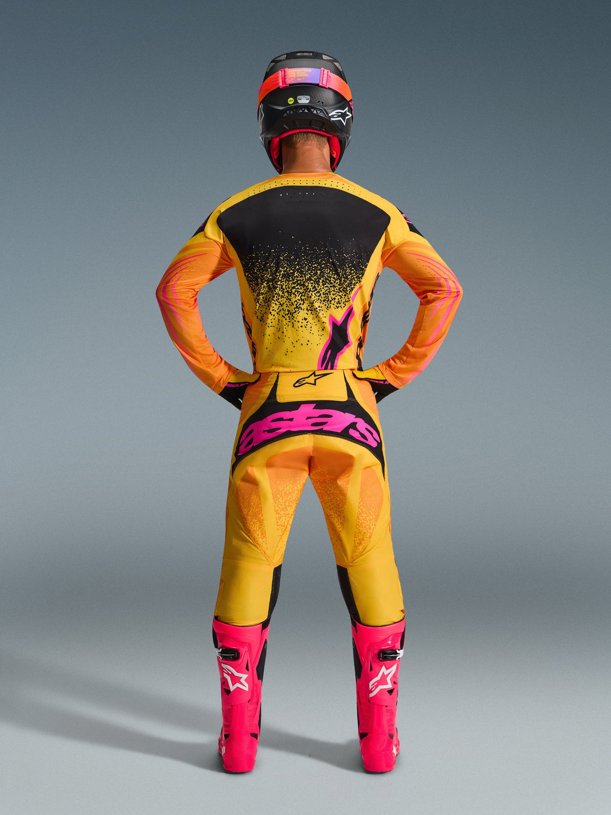 Motocrosskører set bagfra iført Alpinestars 2026 Techstar Nomur bukser i Orange Yellow Pink, med et stort pink logo på sædet, kombineret med en matchende gul og sort jersey, pangfarvede pink støvler og en sort og pink hjelm.