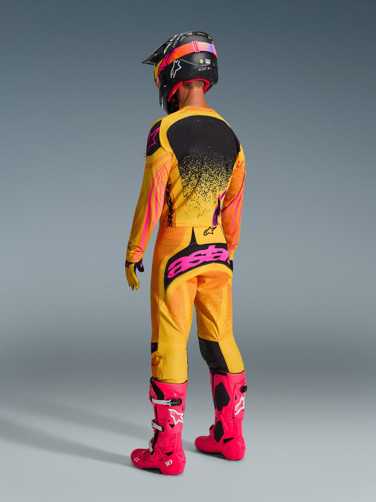 Motocrosskører set bagfra iført komplet racerudstyr, 2026 Techstar Nomur-bukser i Orange Yellow Pink, markant lyserød 'astars'-branding på sædet, matchende trøje, skarpt lyserøde støvler og sort hjelm med lyserøde detaljer, koordineret off-road konkurrenceoutfit.
