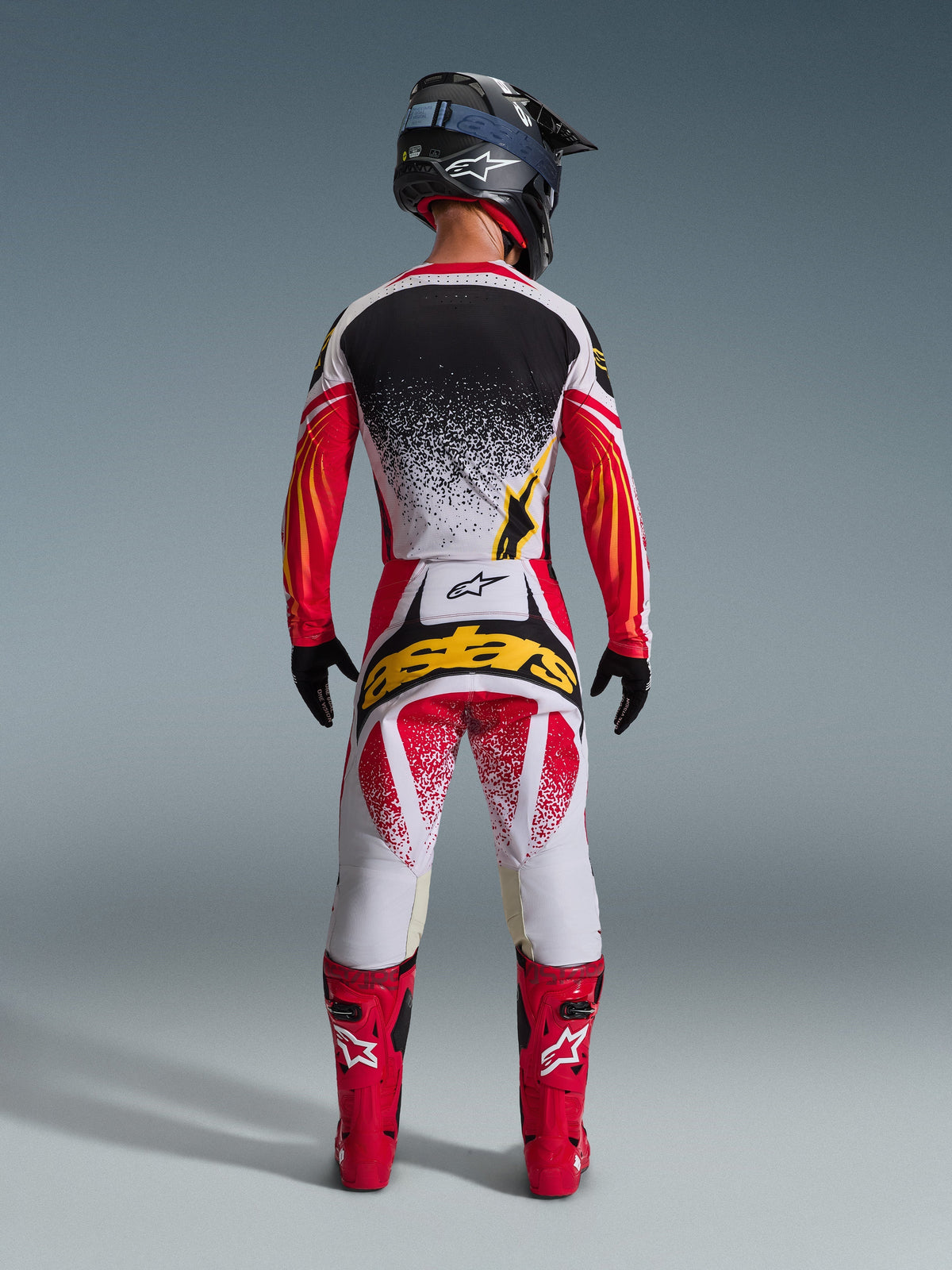 Alpinestars 2026 Techstar Nomur Jersey, set bagfra af en motocross-kører, sort, rød og hvid med gule detaljer, matchende motocross-bukser med iøjnefaldende gul branding, klare røde racerstøvler og en sort hjelm, komplet koordineret udstyrssæt til off-road racing.