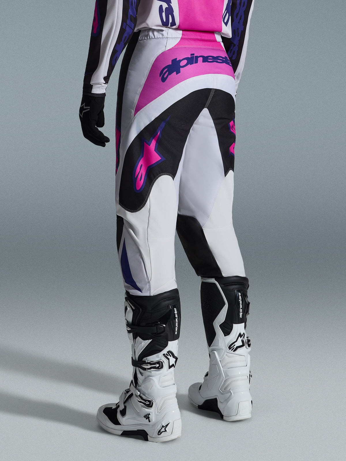 Alpinestars 2026 Fluid Grid bukser, motocross-bukser, sort, lysegrå og lilla, set bagfra af kører iført bukser og hvide og sorte støvler, med lyserøde og lilla Alpinestars-logoer, atletisk pasform til off-road racing