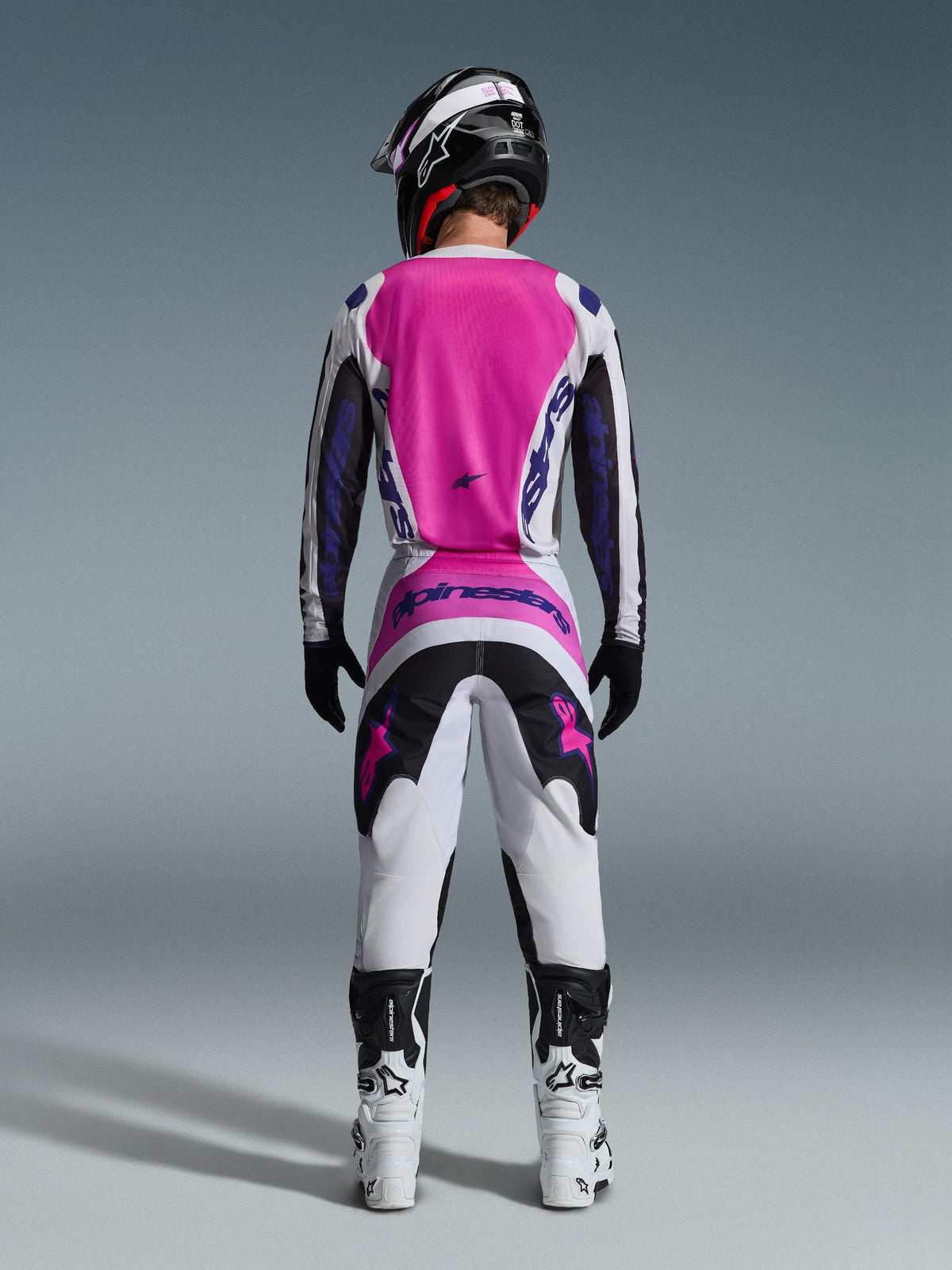 Motocross-kører set bagfra iført Alpinestars 2026 Fluid Grid-bukser og matchende trøje, pink, hvid og sort med lilla branding, sort hjelm, hvide og sorte støvler, koordineret racersæt til off-road motocross