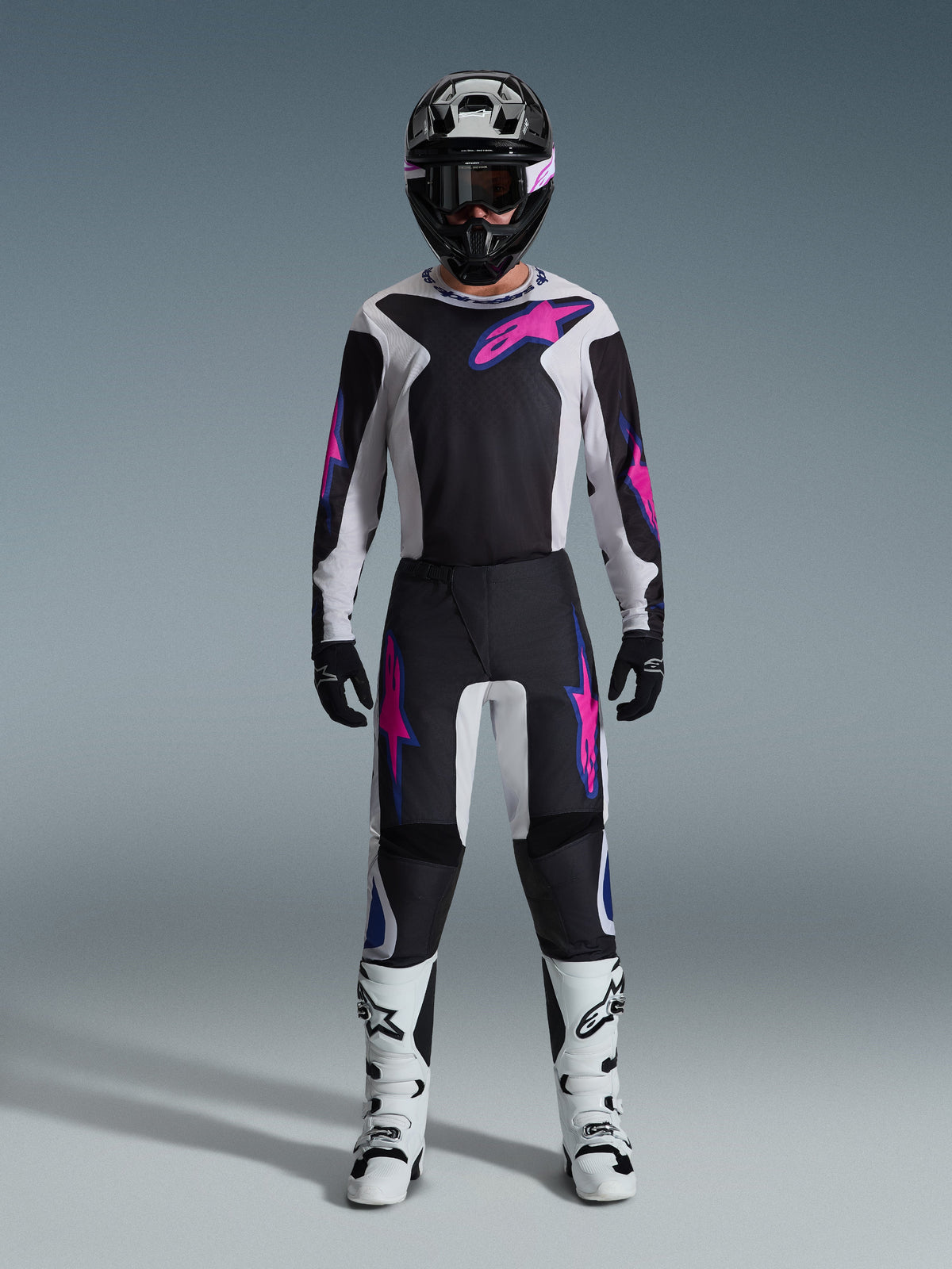 Motocross-kører iført Alpinestars 2026 Fluid Grid-udstyrssæt, sort og lysegrå trøje med skarpe lyserøde logoer, sorte og hvide bukser med lyserød og blå grafik, sort hjelm med lyserøde briller, hvide og sorte støvler, komplet off-road-racerudstyr