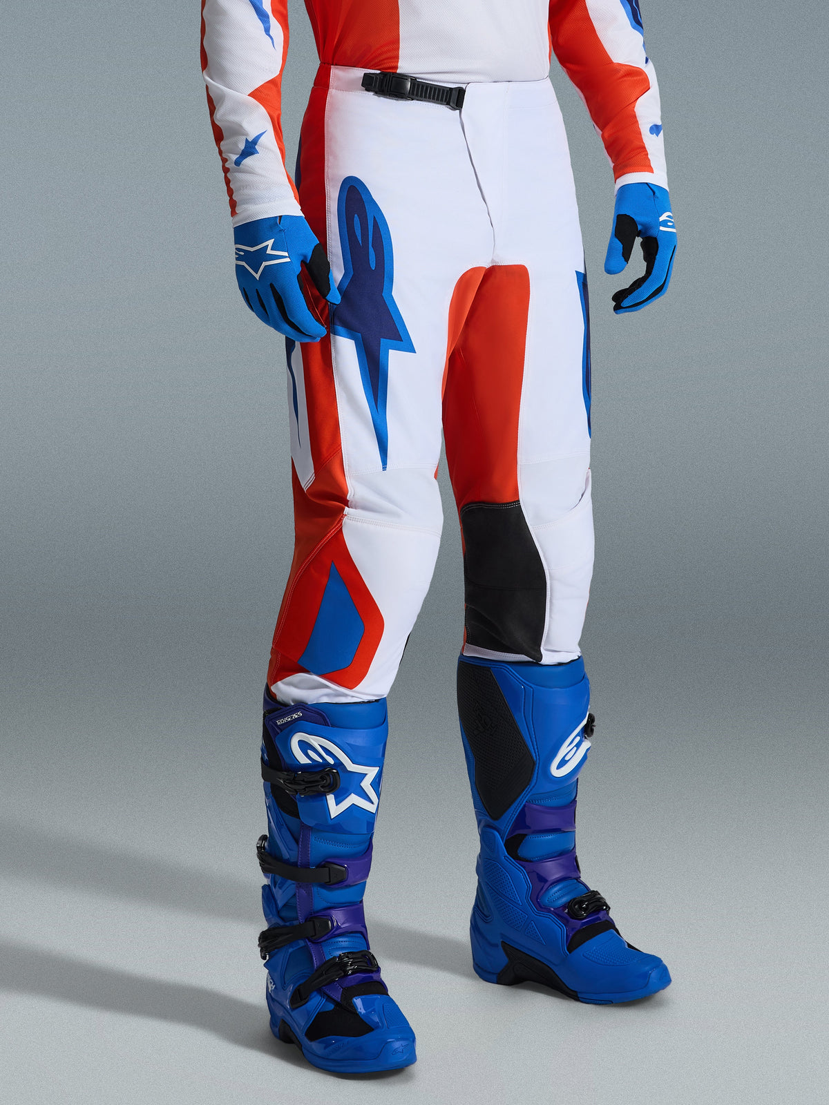 Alpinestars 2026 Fluid Grid bukser, motocross-bukser, orange og UCLA-blå, båret med matchende blå handsker og støvler, hvid base med markant grafisk branding, sorte indvendige knæpaneler og et ergonomisk design til off-road kørsel.