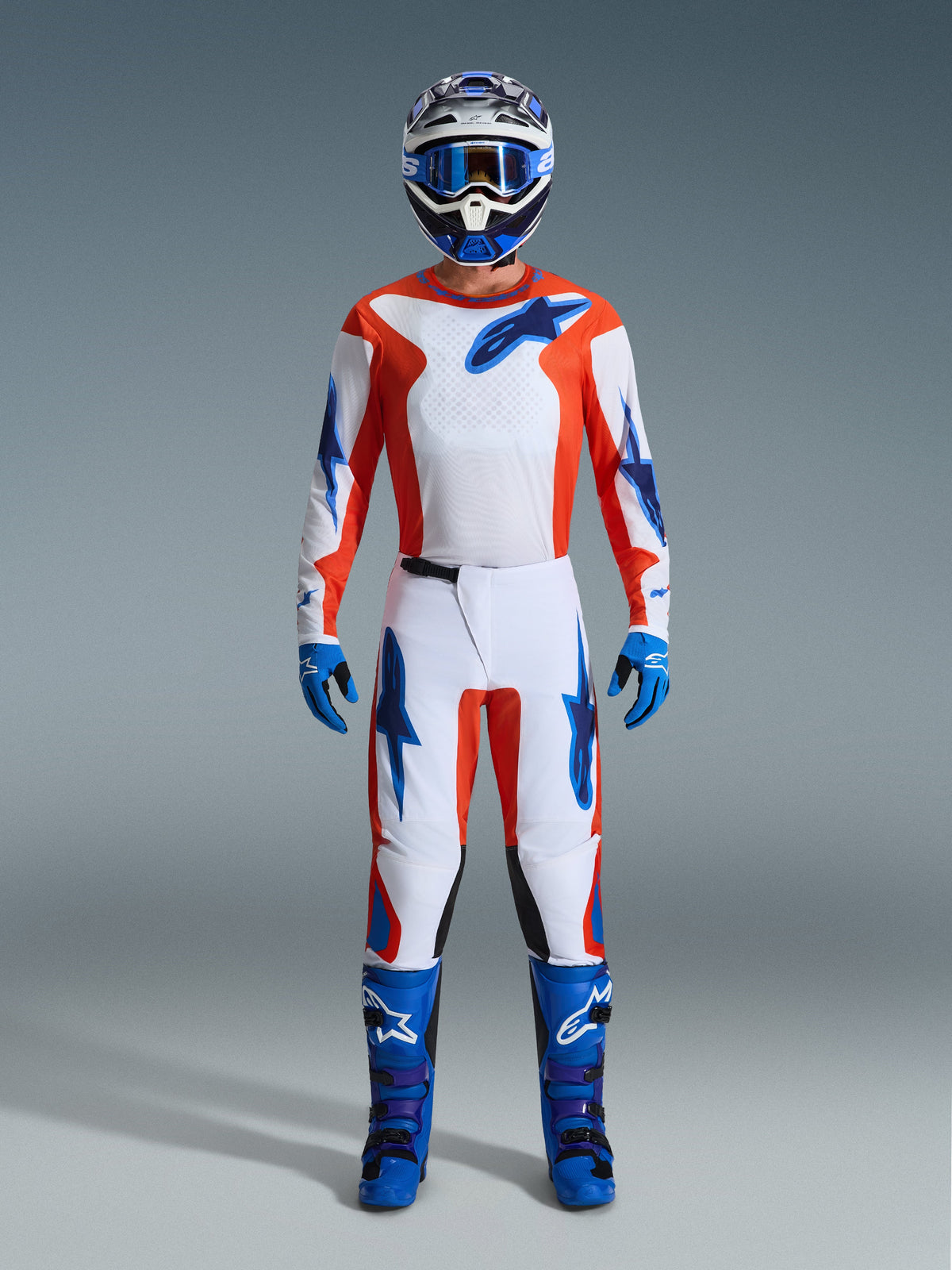 Motocross-kører iført komplet racerudstyr, Alpinestars 2026 Fluid Grid-bukser i Orange og UCLA Blue, hvid jersey med orange ærmer og blå logoer, matchende blå hjelm og goggles, blå støvler og blå handsker, koordineret outfit til off-road kørsel