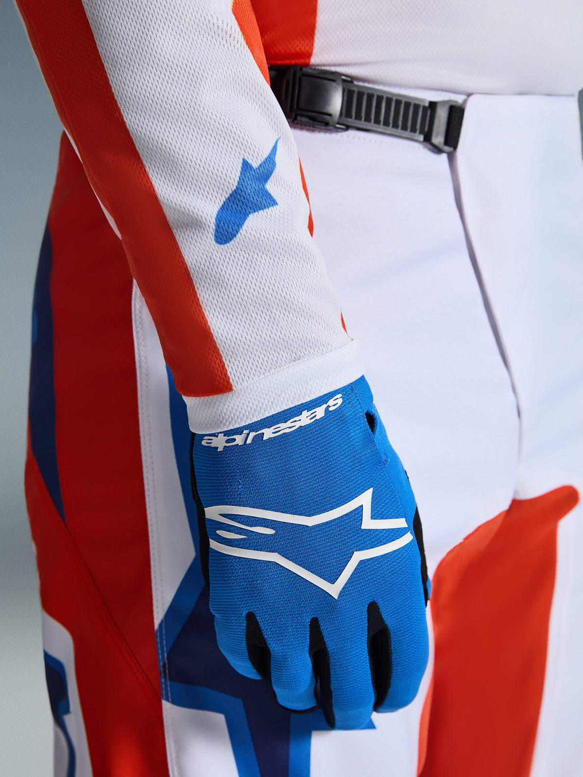 Alpinestars 2026 Fluid Grid bukser, motocross-bukser, orange og UCLA-blå, nærbillede med en matchende blå handske og hvidt mesh-jerseyærme med blå logoer, viser skraldelukningen i taljen og den atletiske pasform.