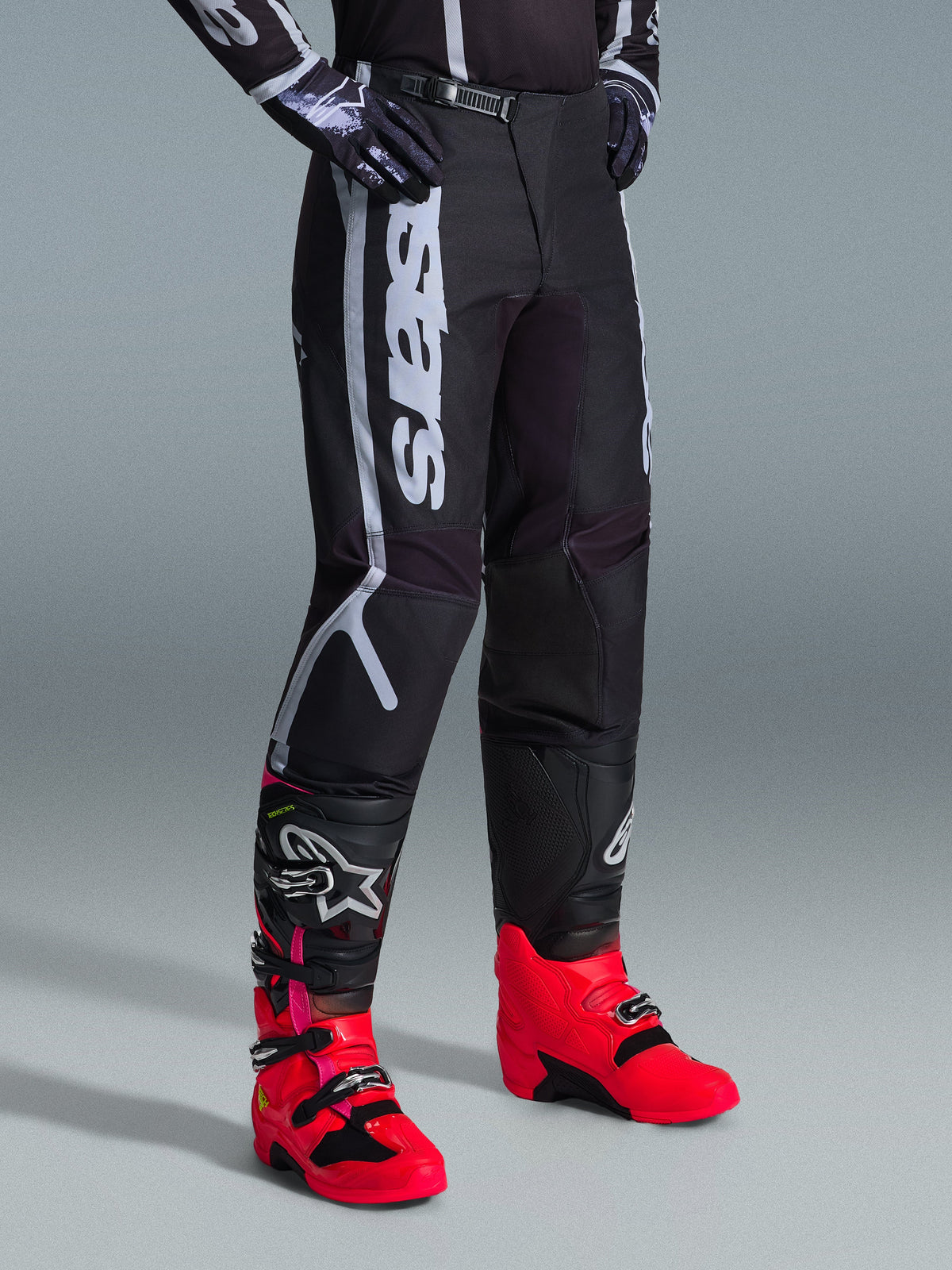 Alpinestars 2026 Fluid Apex bukser, motocross-bukser, sort og grå, båret med livlige pink og røde støvler og matchende sort og hvid trøje, med markant lodret Alpinestars-branding på låret, forstærkede paneler og skraldelukning i taljen for off-road ydeevne.