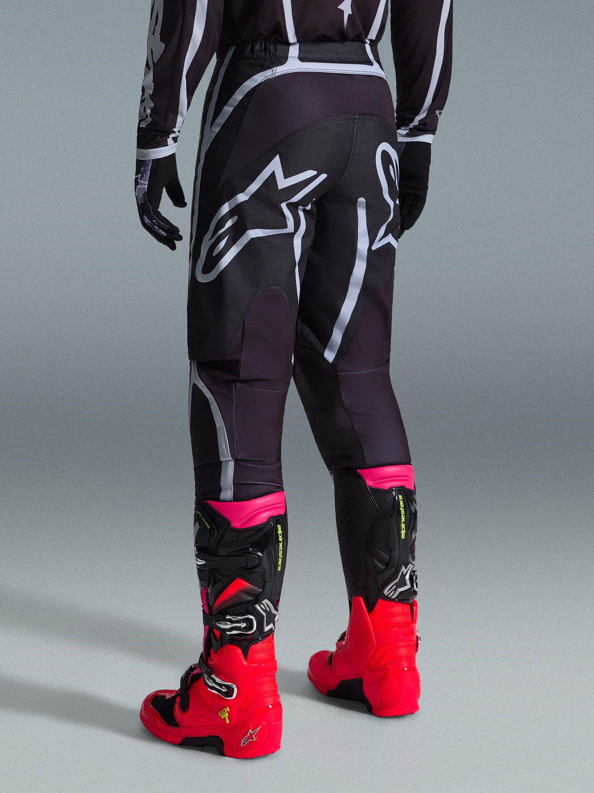 Alpinestars 2026 Fluid Apex bukser, motocrossbukser, sort og grå, set bagfra med store hvide Alpinestars-logoer på sædet, båret med stærkt røde og lyserøde støvler, holdbar 600D polystof-konstruktion til off-road kørsel