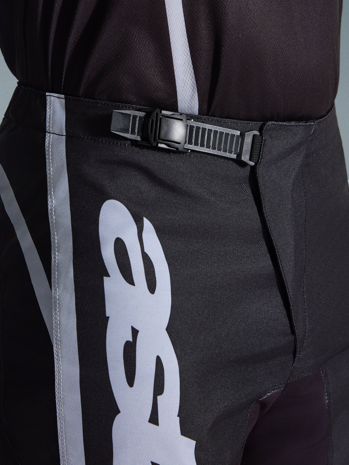 Alpinestars 2026 Fluid Apex bukser, motocrossbukser, sort og grå, nærbillede af det integrerede skraldelukningssystem for en sikker pasform, med hvid logografik på slidstærkt 600D polystof.