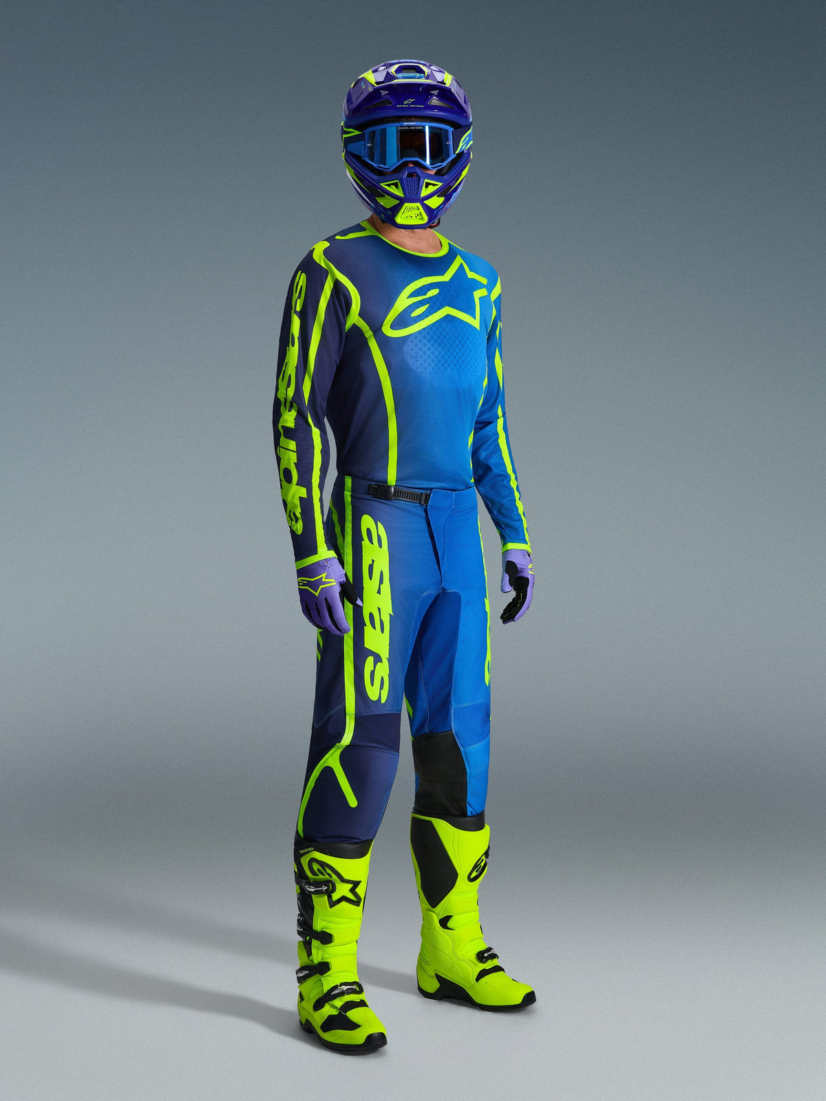 Motocrosskører iført Alpinestars 2026 Fluid Apex-bukser i blå og gul fluo, koordineret med en matchende trøje, blå og gul hjelm og klare gule støvler, professionelt off-road-udstyrssæt til motocross.