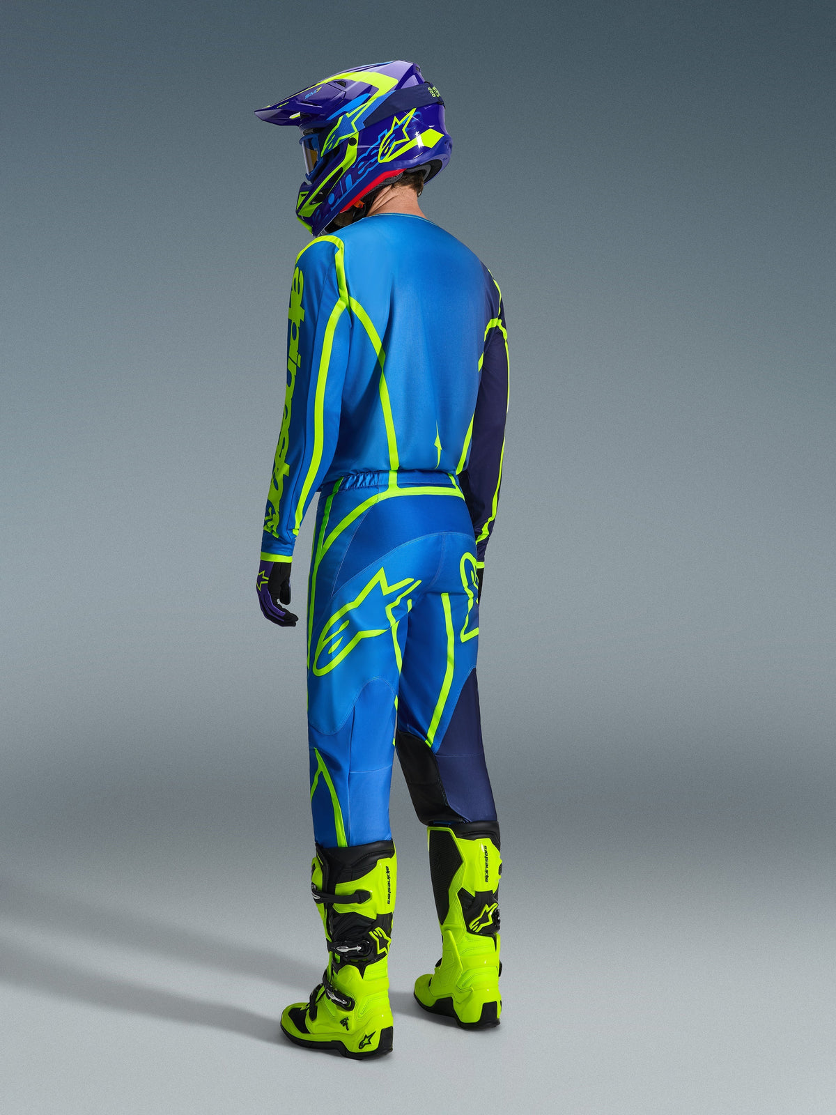 Motocross-kører set bagfra iført 2026 Alpinestars Fluid Apex bukser i blå og neon-gul, matchende blå og gul trøje og hjelm, samt klare neon-gule støvler, koordineret racerudstyr til off-road kørsel.