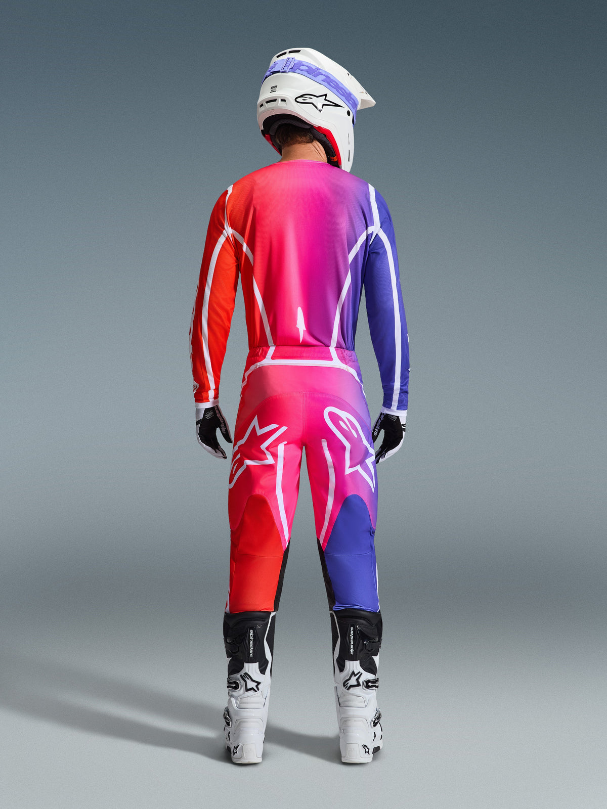 Alpinestars 2026 Fluid Apex-bukser og matchende trøje, motocross-kører set bagfra, levende rød, pink og lilla farveovergang, hvid hjelm og hvide støvler, komplet koordineret udstyrssæt til off-road racing
