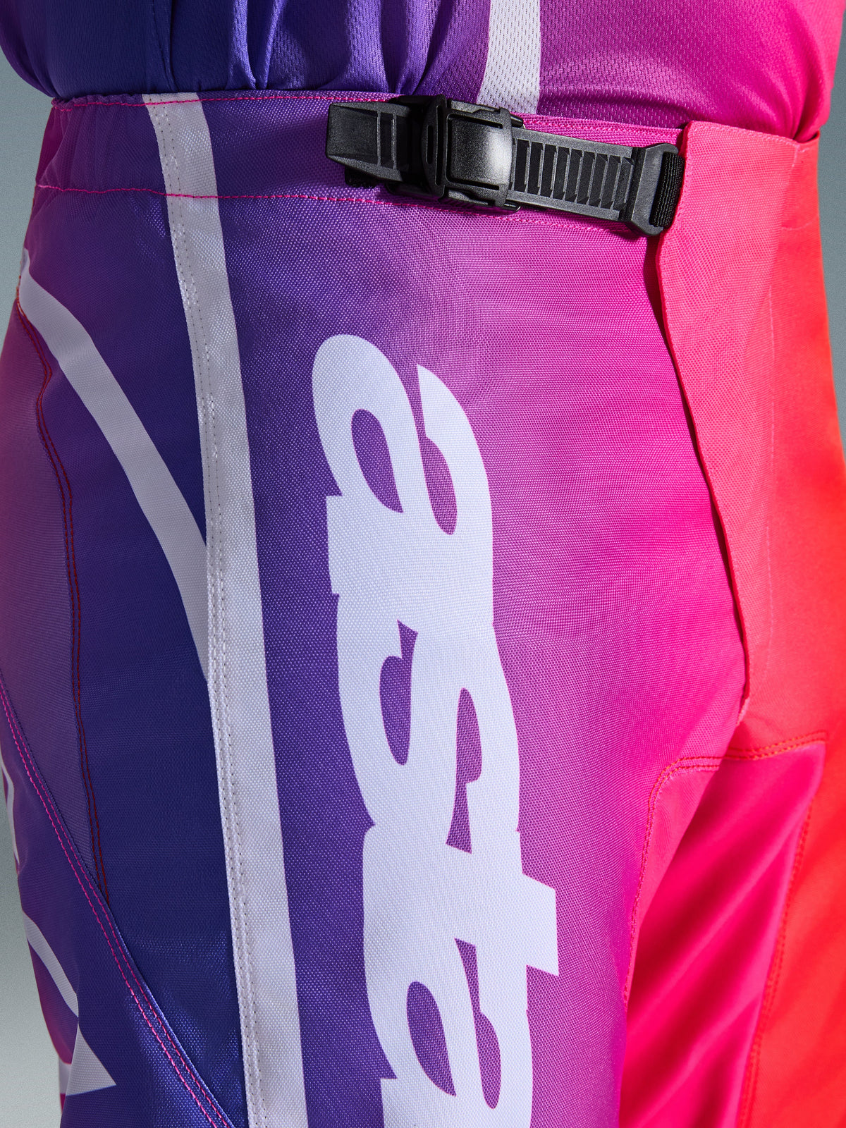Alpinestars 2026 Fluid Apex bukser, motocross-bukser, multifarvet lilla, pink og rød, tætbillede af talje- og hofteområdet med et sort skraldelukningssystem og stort hvidt lodret Alpinestars-logo, levende gradient-design med holdbar polystof-tekstur