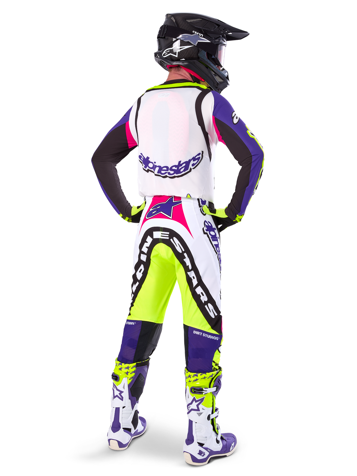 Motocross-kører iført Alpinestars Limited Edition Supertech Dirt Studios® bukser i hvid, lilla og neon-gul, set bagfra med matchende trøje, støvler og sort carbon-hjelm, professionelt off-road racerudstyr med markant grafik og neon-detaljer.