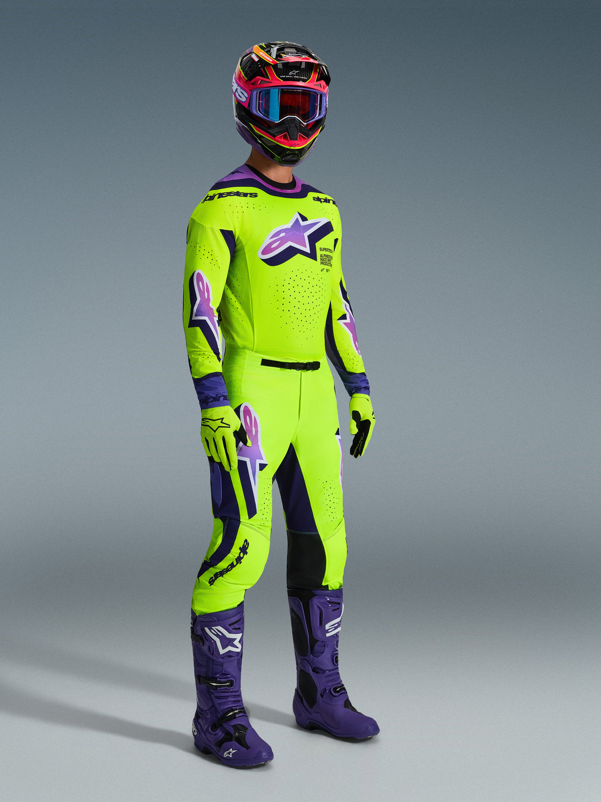 Alpinestars 2026 Supertech Vista udstyrssæt, professionel motocross-kører iført en stærk limegrøn trøje og bukser med lilla detaljer og logoer, matchende limegrønne handsker, lilla støvler og en flerfarvet hjelm med blå briller, koordineret atletisk udstyr til off-road racing