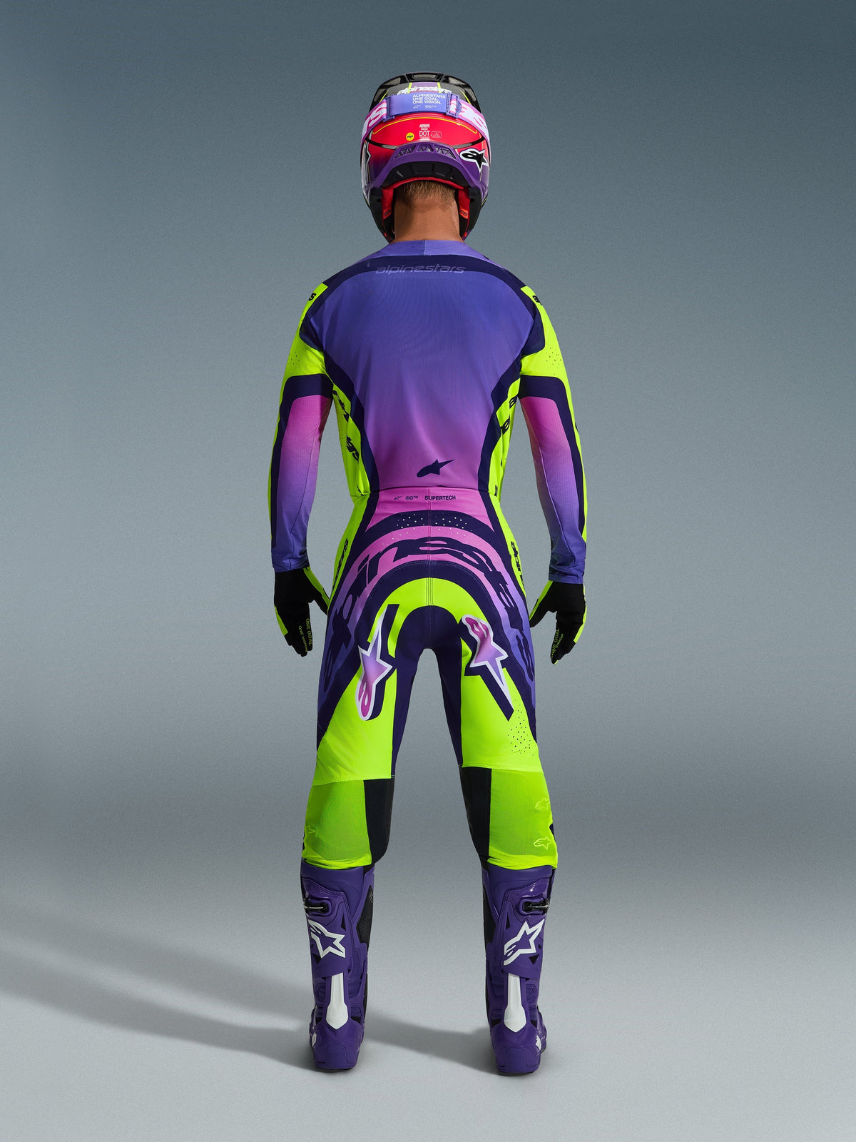 Motocross-kører set bagfra iført 2026 Supertech Vista-trøje og matchende bukser, livligt gult og lilla design med lyserøde gradienter, lilla støvler med hvide Alpinestars-logoer, sorte handsker og en rød og lilla racerhjelm, professionelt racersæt.