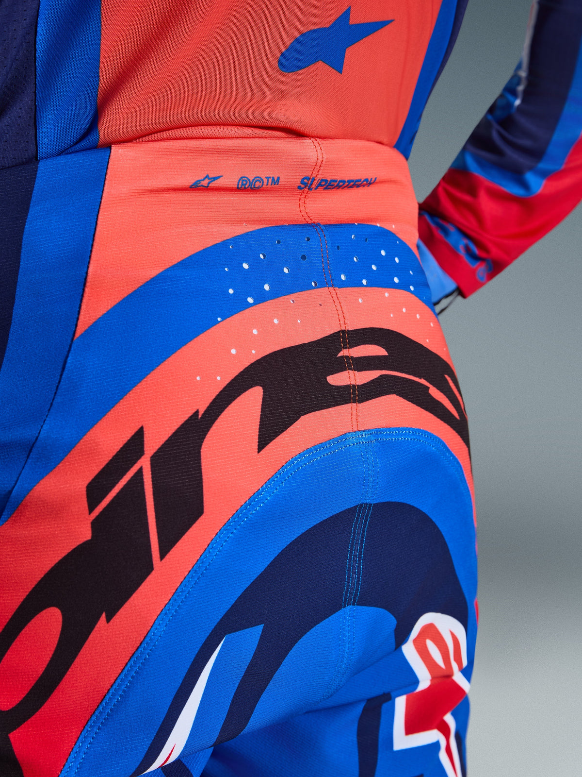 Alpinestars 2026 Supertech Vista bukser, motocrossbukser, mørkeblå, Hot Coral og koboltblå, nærbillede bagfra der viser markant branding, laserskårne perforeringer til ventilation og 'SUPERTECH'-logo på linningen, performance-strækmateriale til off-road racing