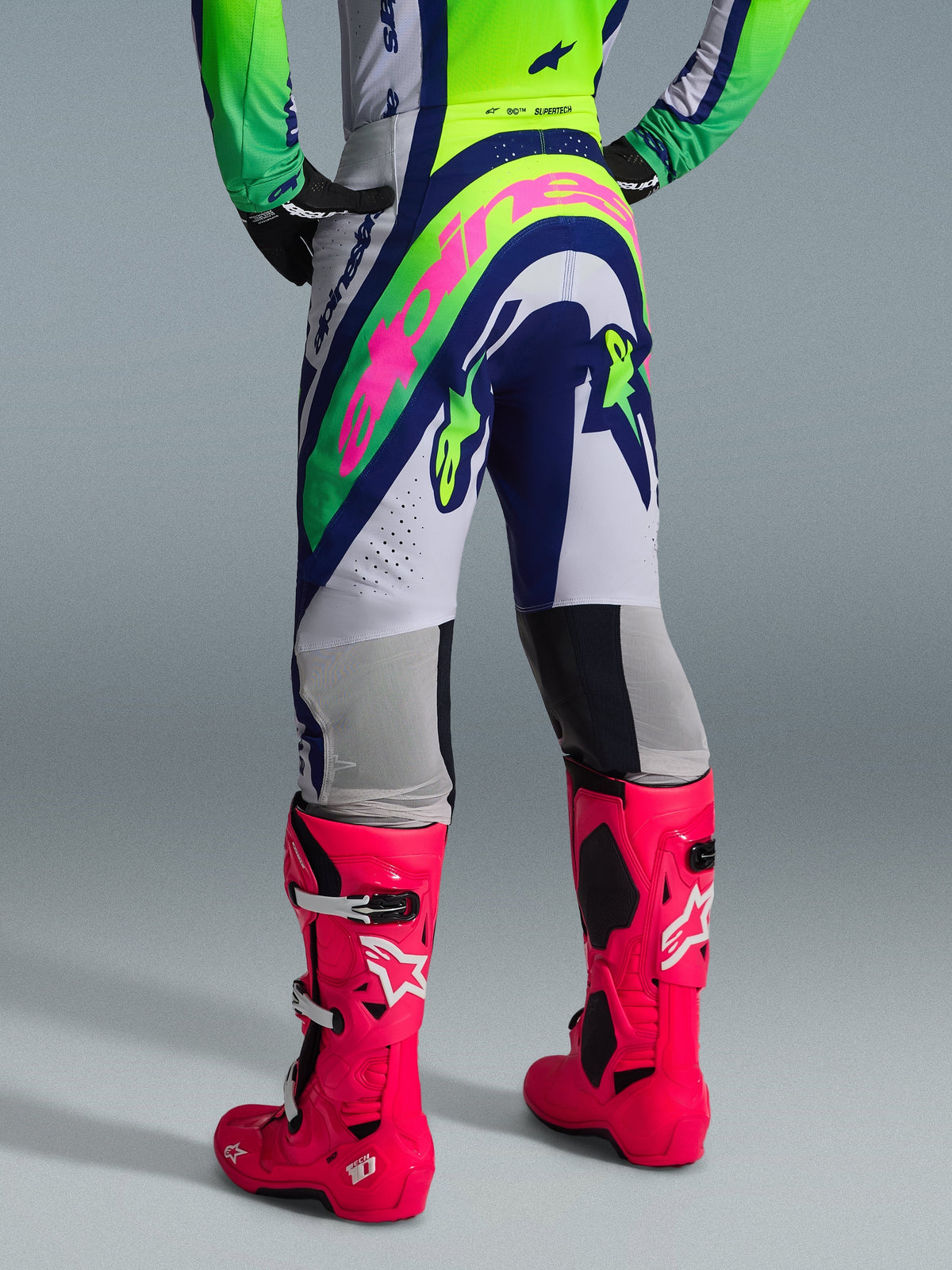Alpinestars 2026 Supertech Vista bukser, motocrossbukser, lysegrå, neongul og blå, set bagfra med et stort lyserødt Alpinestars-logo, båret med matchende racingtrøje og stærkt lyserøde Tech 10-støvler, professionelt terrænudstyr