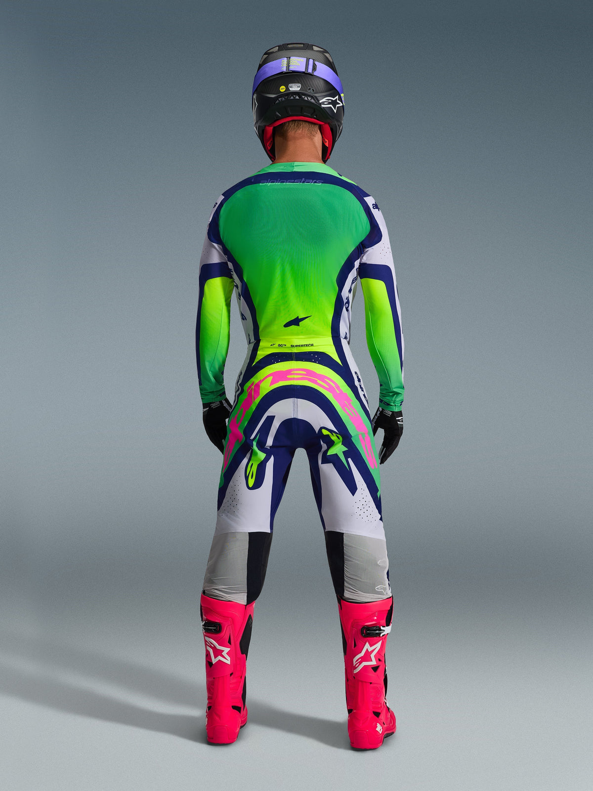 Alpinestars 2026 Supertech Vista bukser, motocrossbukser, lysegrå, gul fluo og blå, set bagfra af kører iført matchende jersey med grøn og gul farveovergang og skarpe lyserøde støvler, med markant lyserødt Alpinestars-brand på bagdelen, professionel atletisk pasform til terrænkørsel