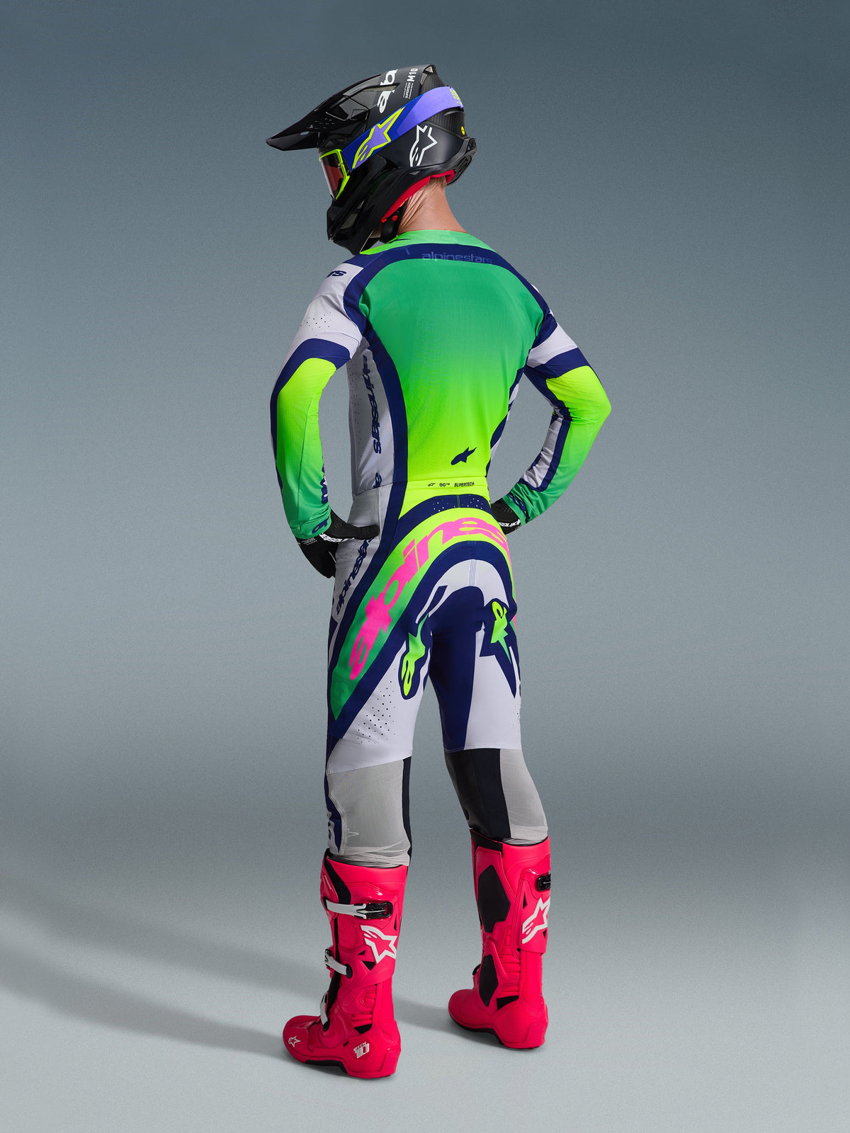 Set bagfra af en motocross-kører iført Alpinestars 2026 Supertech Vista Gear Set, hvid og limegrøn trøje med blå detaljer, matchende bukser med et iøjnefaldende pink logo hen over ryggen, lysende pink racerstøvler og en sort hjelm med limegrønne briller, professionelt terrænkørselstøj