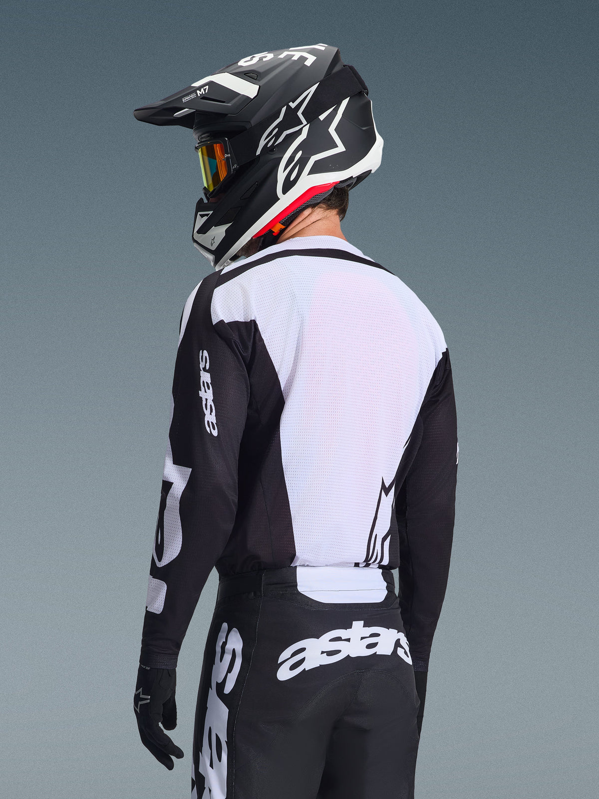 Motocross rider from behind wearing the Alpinestars 2026 Racer Air Riway Jersey, Black and White, featuring a white breathable mesh back panel and black sleeves with 'astars' branding, matching black pants with white logos, black helmet with white accents, professional off-road gear for high-temperature performance.