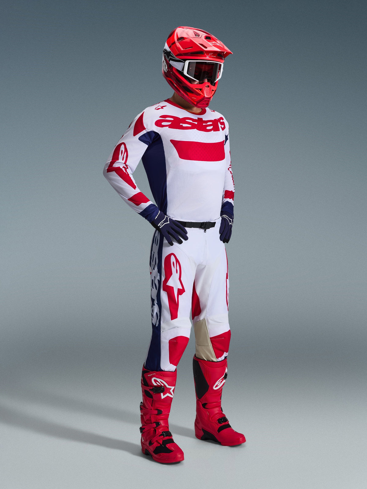 Motocrosskører iført Alpinestars 2026 Racer Riway-udstyrssæt, hvid trøje med røde og blå detaljer og 'astars'-branding, matchende hvide bukser med store røde logoer og mørkeblå sidepaneler, rød hjelm, røde og sorte støvler og mørkeblå handsker, professionelt racersæt til terrænkørsel.