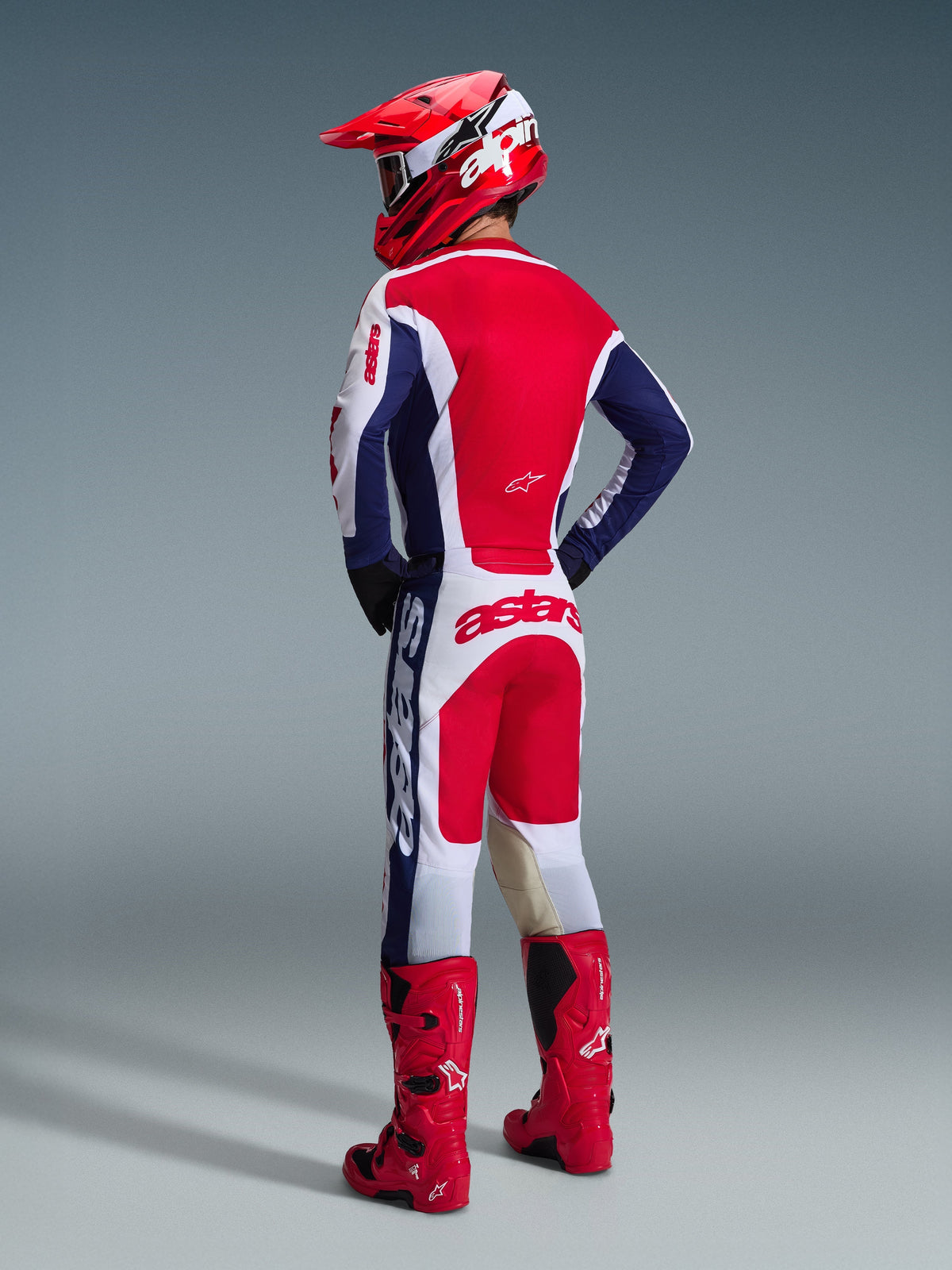 Motocrosskører set bagfra iført Alpinestars 2026 Racer Riway-udstyrssættet i hvid, blå og rød, inklusiv en langærmet trøje med Astars-branding og matchende bukser, koordineret med en rød hjelm og røde off-road støvler.