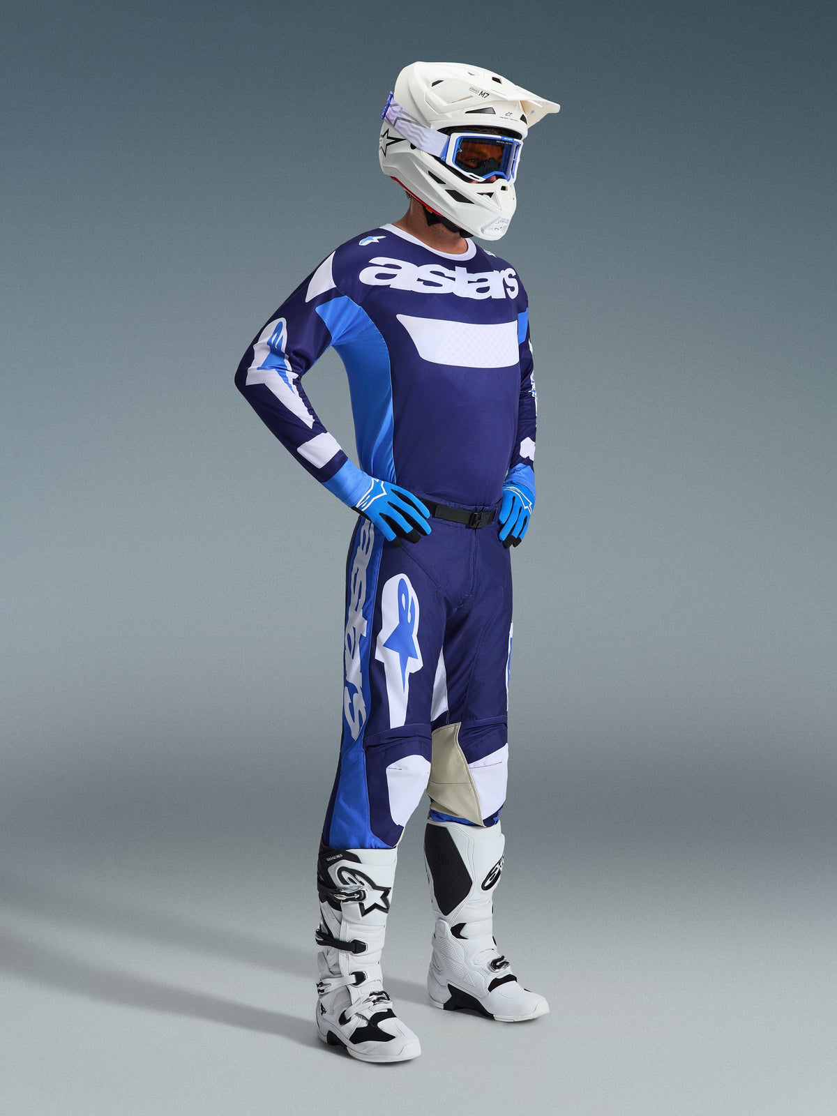 Motocrosskører iført Alpinestars 2026 Racer Riway-udstyrssæt i UCLA Blue og hvid, bestående af en blå og hvid trøje med 'astars'-branding, matchende bukser, en hvid hjelm med blå goggles og hvide off-road støvler, komplet raceroutfit til motocross.