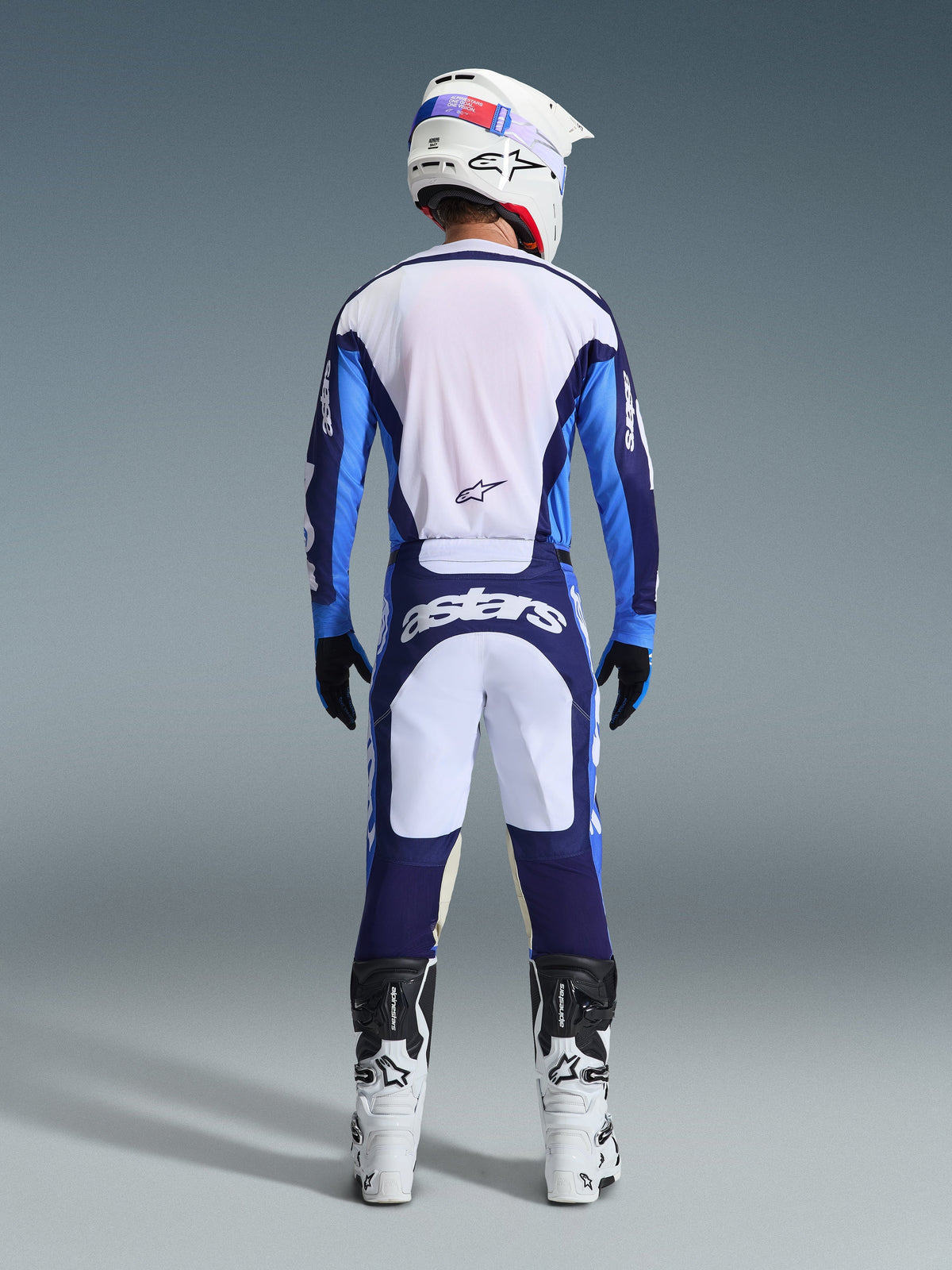 Set bagfra af en motocrosskører iført Alpinestars 2026 Racer Riway Jersey og bukser i Ucla Blue og hvid, koordineret med en hvid hjelm, sorte handsker og hvide motocrossstøvler, højpræstationsudstyr til off-road racing