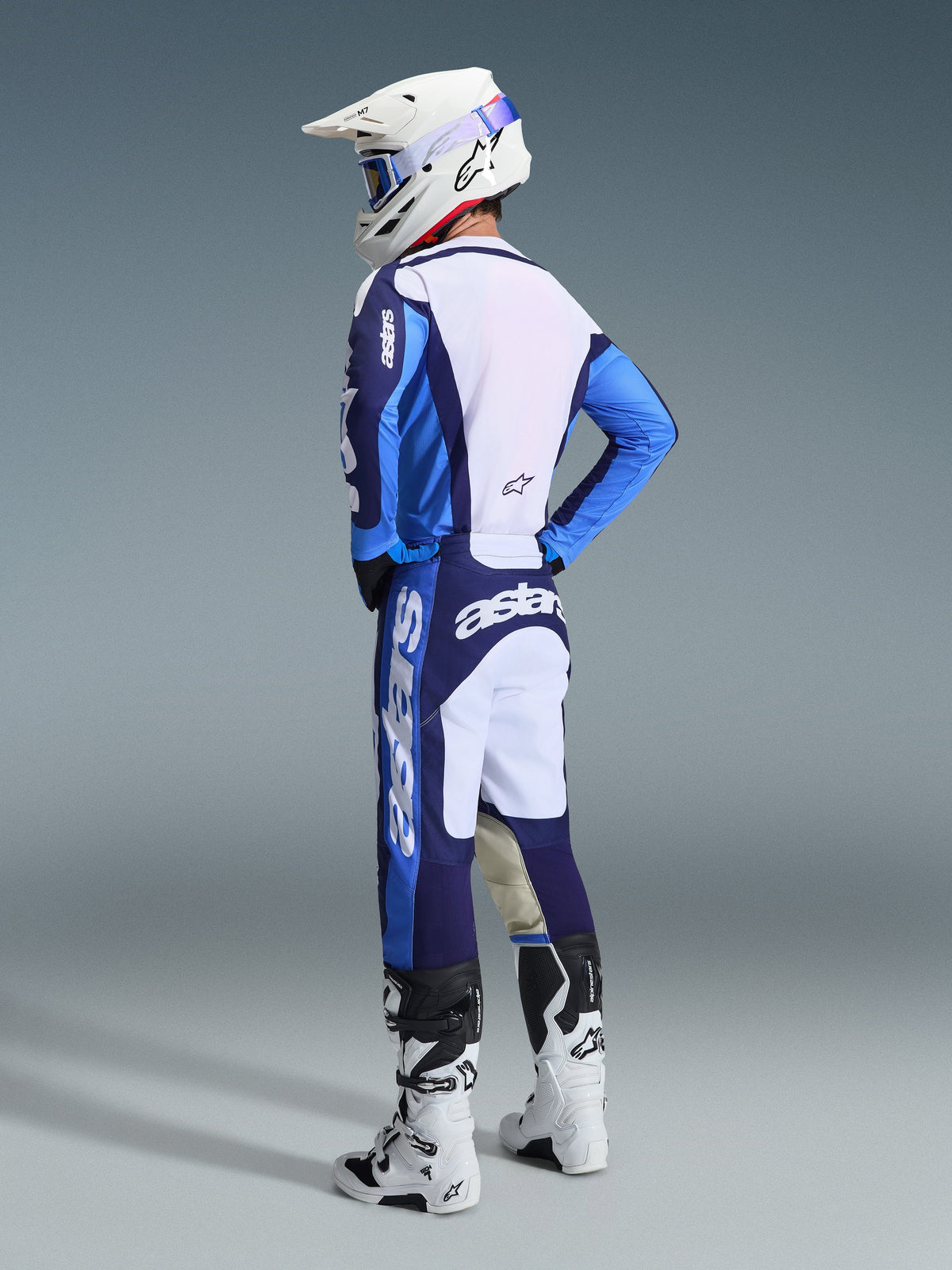 Alpinestars 2026 Racer Riway-udstyrssæt i UCLA Blue og hvid, motocrosskører set bagfra fra siden, blå og hvid jersey med 'astars'-branding, matchende blå og hvide bukser, hvid motocrosshjelm med blå goggles og hvide og sorte Tech-serie støvler.