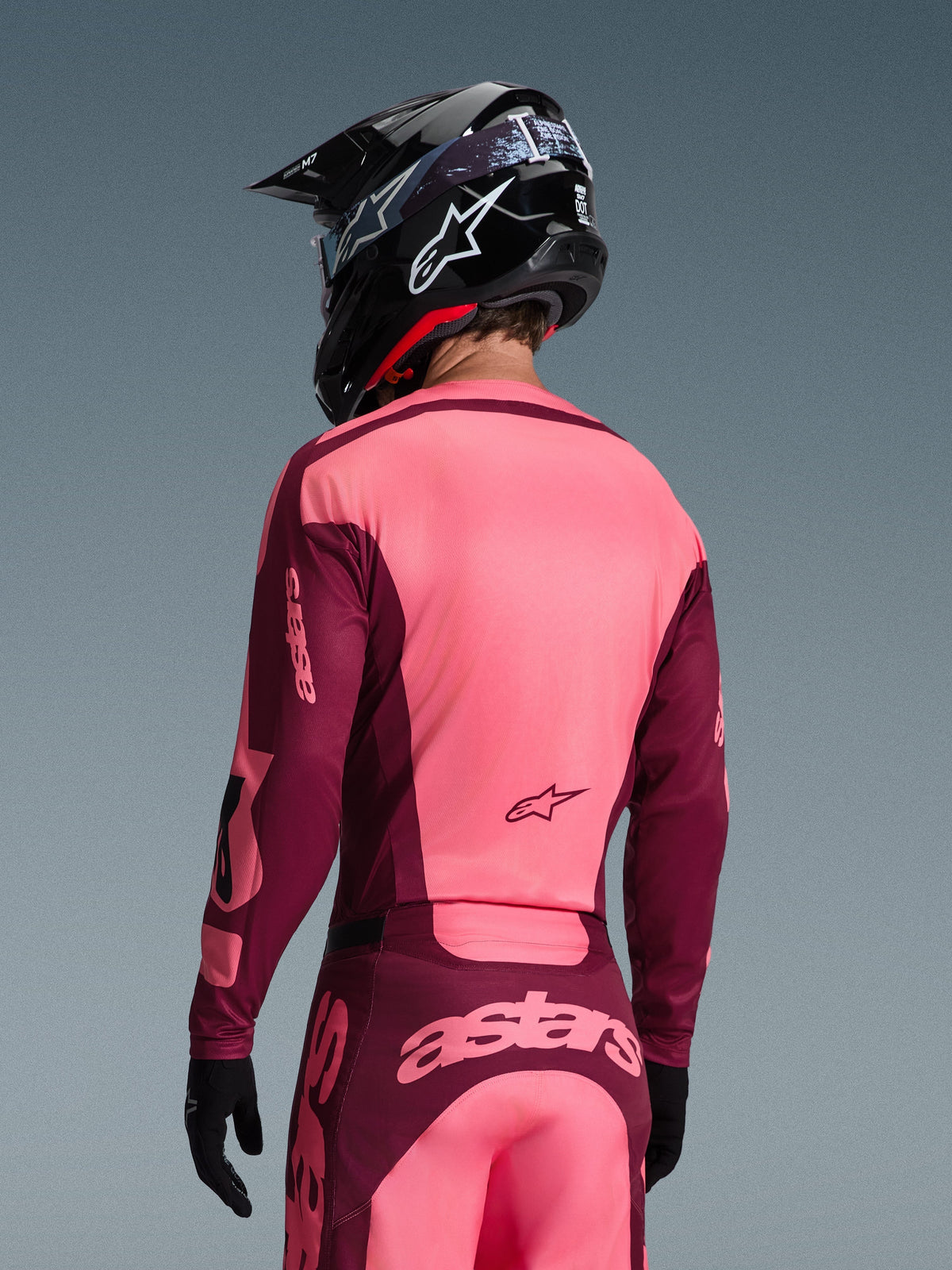 Motocrosskører set bagfra iført Alpinestars 2026 Racer Riway-trøje og matchende bukser, Dark Red og Coral farvekombination, sort hjelm med hvidt Alpinestars-logo, sorte handsker, professionelt koordineret udstyr til off-road kørsel